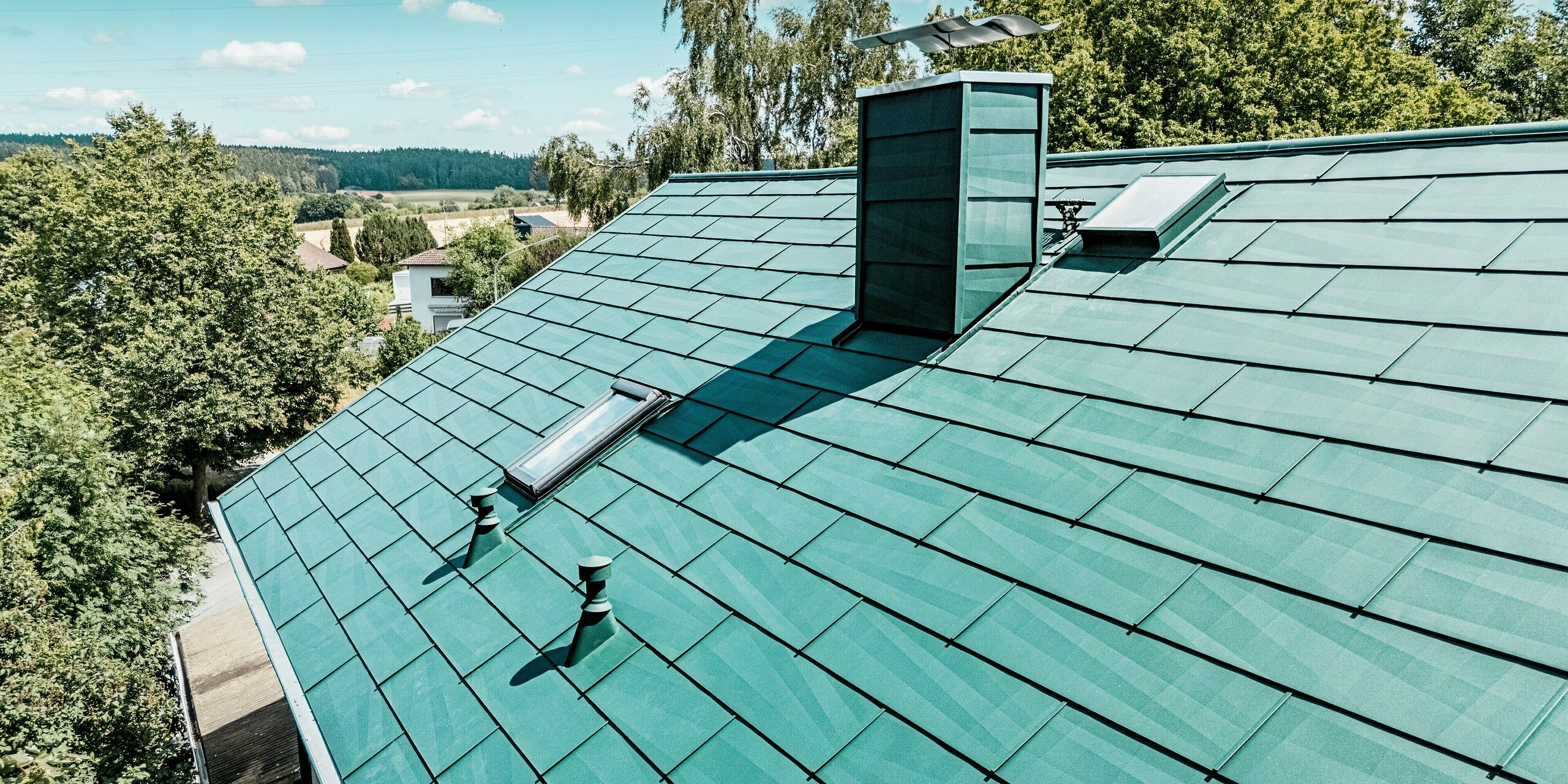 Toiture modernisée d'une maison individuelle à Schwarzenbach-Martinlamitz, réalisée avec le panneau de toiture durable PREFA FX.12 en vert mousse P.10. L'image détaillée met en valeur cette toiture en aluminium aux lignes épurées, à la surface texturée, aux puits de lumière intégrés et à la cheminée élégamment revêtue. Le jeu de lumière se reflétant sur les panneaux accentue le design moderne. Grâce à sa surface en aluminium robuste, la toiture offre une protection optimale contre les intempéries, ne nécessite aucun entretien et arbore une esthétique élégante et intemporelle.