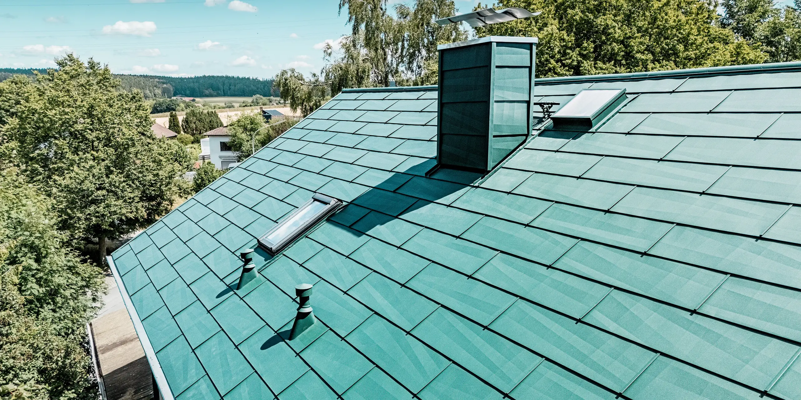 Toiture modernisée d'une maison individuelle à Schwarzenbach-Martinlamitz, réalisée avec le panneau de toiture durable PREFA FX.12 en vert mousse P.10. L'image détaillée met en valeur cette toiture en aluminium aux lignes épurées, à la surface texturée, aux puits de lumière intégrés et à la cheminée élégamment revêtue. Le jeu de lumière se reflétant sur les panneaux accentue le design moderne. Grâce à sa surface en aluminium robuste, la toiture offre une protection optimale contre les intempéries, ne nécessite aucun entretien et arbore une esthétique élégante et intemporelle.