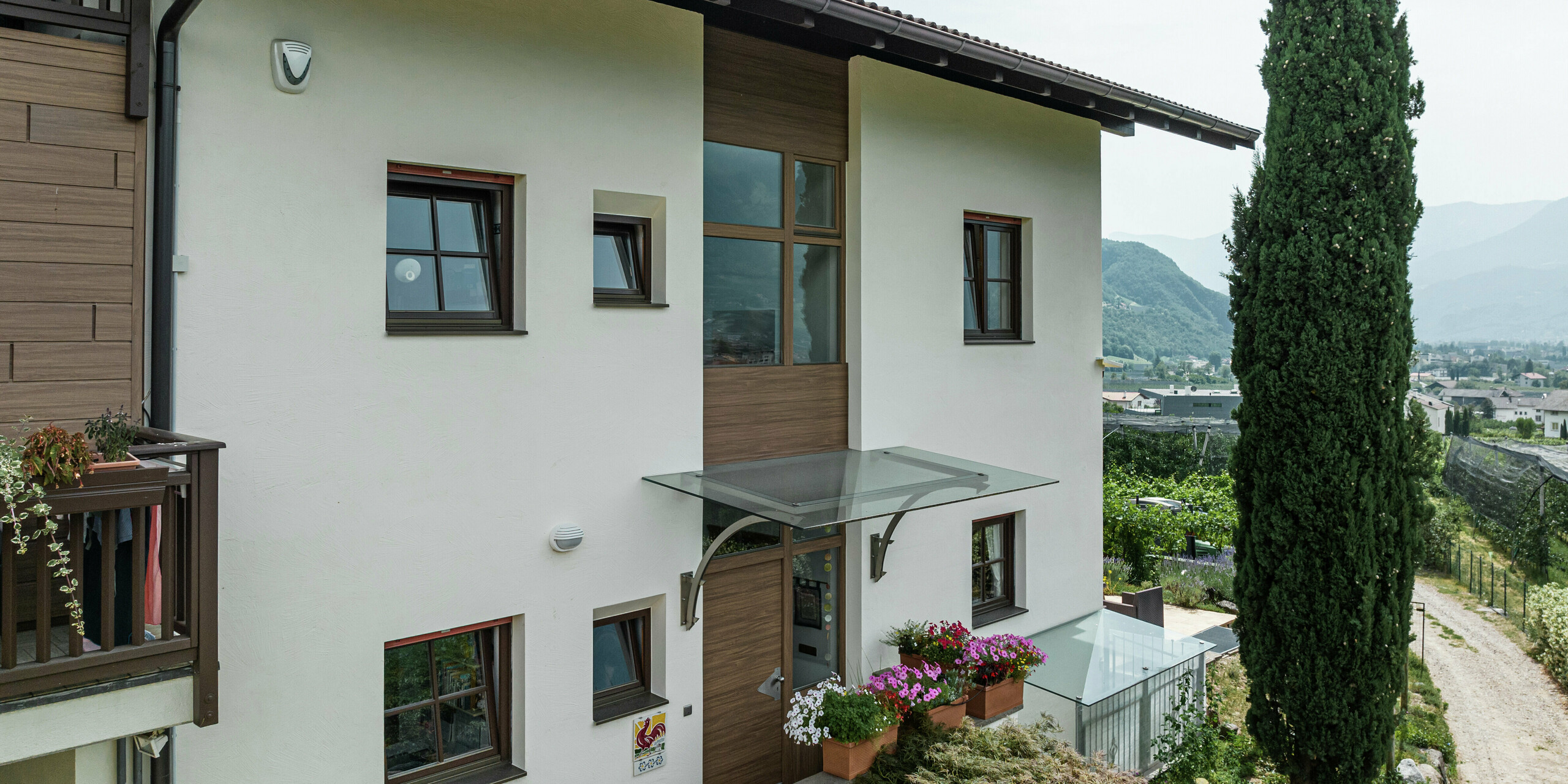 Zone d'entrée des appartements de vacances Prechtlhof à Merano, dans le Tyrol du Sud, avec une façade moderne en plâtre et des Sidings PREFA en noyer foncé. La façade en aluminium sans entretien avec un aspect bois complète le design traditionnel et garantit une solution durable et résistante aux intempéries.