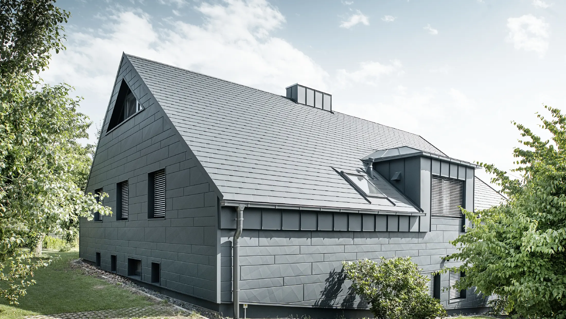 Grande maison individuelle avec toit en pente et nombreuses fenêtres. L'ensemble de la maison a été revêtu d'une enveloppe en aluminium anthracite, la toiture a été réalisée avec des bardeaux en aluminium PREFA et la façade avec du Siding.X anthracite.