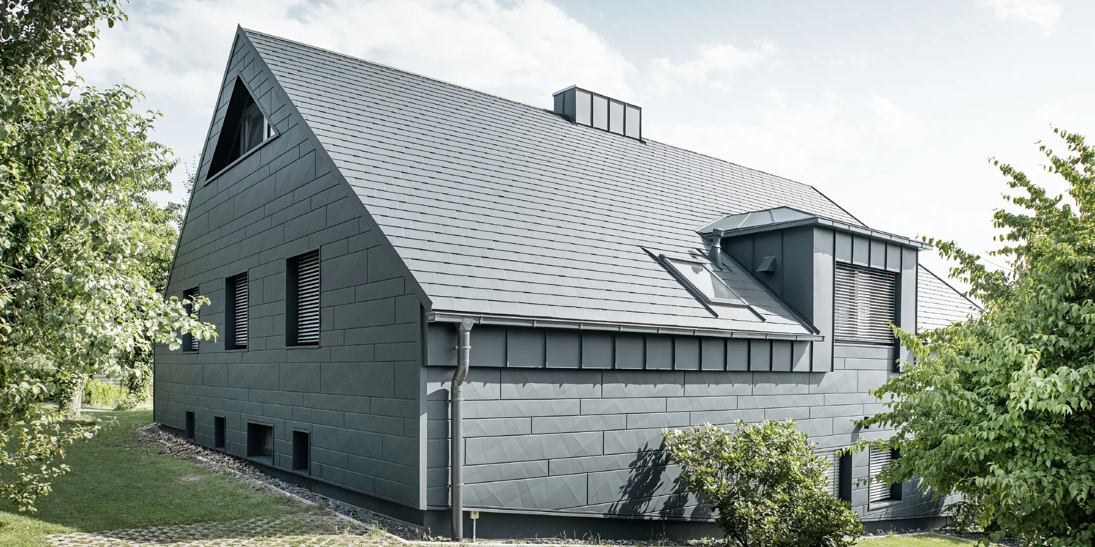 Grande maison individuelle avec toit en pente et nombreuses fenêtres. L'ensemble de la maison a été revêtu d'une enveloppe en aluminium anthracite, la toiture a été réalisée avec des bardeaux en aluminium PREFA et la façade avec du Siding.X anthracite.