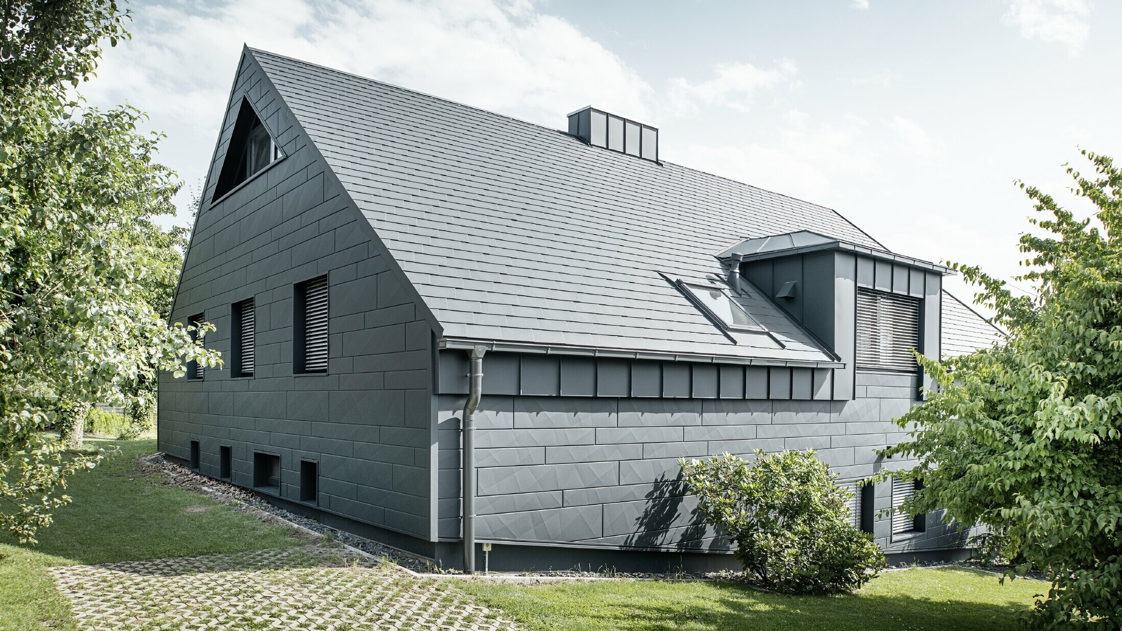 Grande maison individuelle avec toit en pente et nombreuses fenêtres. L'ensemble de la maison a été revêtu d'une enveloppe en aluminium anthracite, la toiture a été réalisée avec des bardeaux en aluminium PREFA et la façade avec du Siding.X anthracite.