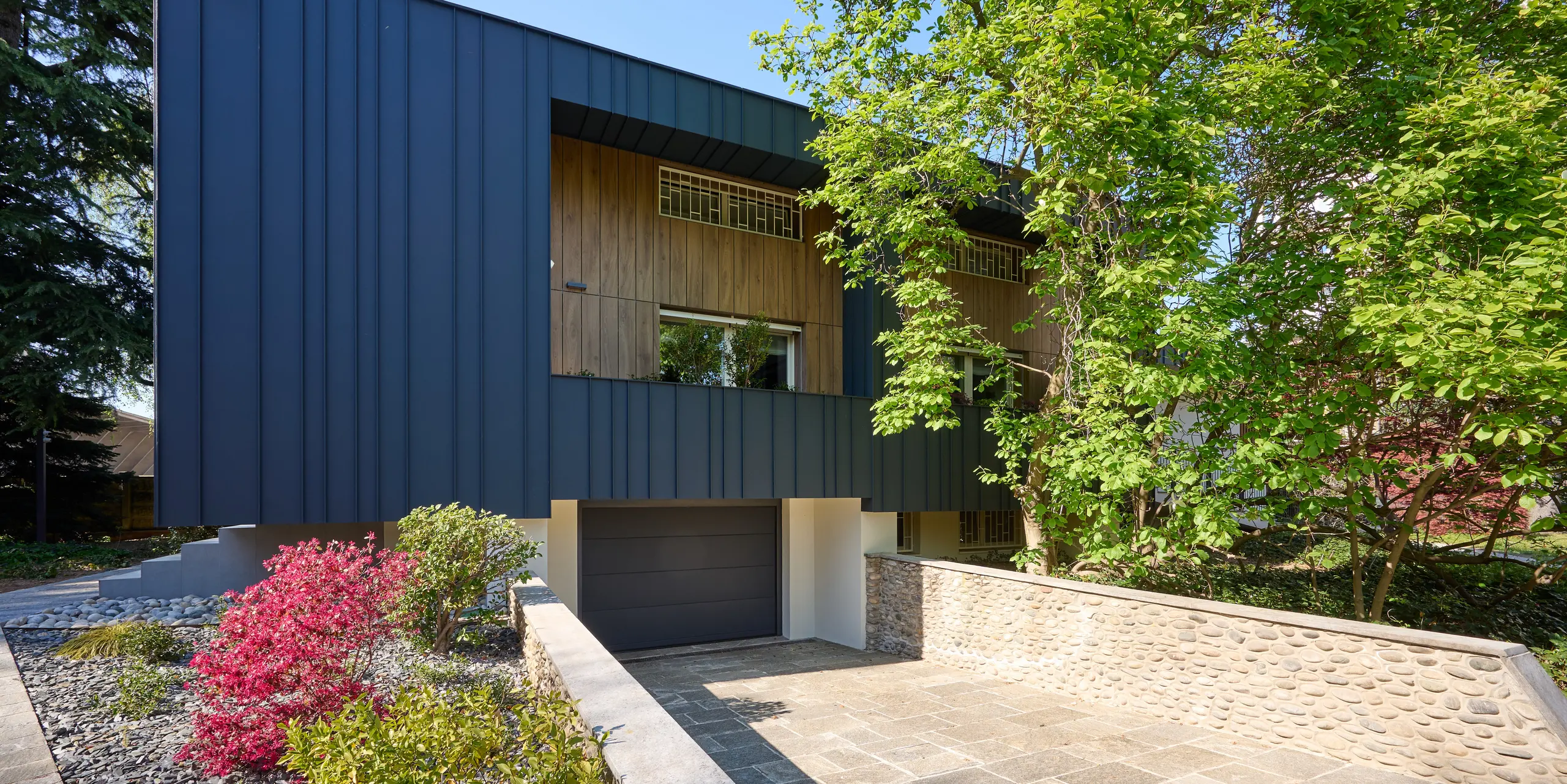 Vue de face de la Villa Castellanza, revêtue d'une élégante façade PREFALZ couleur anthracite P.10. Les joints debout apparents créent un contraste saisissant avec les éléments en bois chaleureux et le jardin luxuriant. Le bardage en aluminium PREFA, de haute qualité, séduit par ses lignes architecturales épurées, sa grande résistance aux intempéries, sa durabilité et son esthétique résolument moderne.