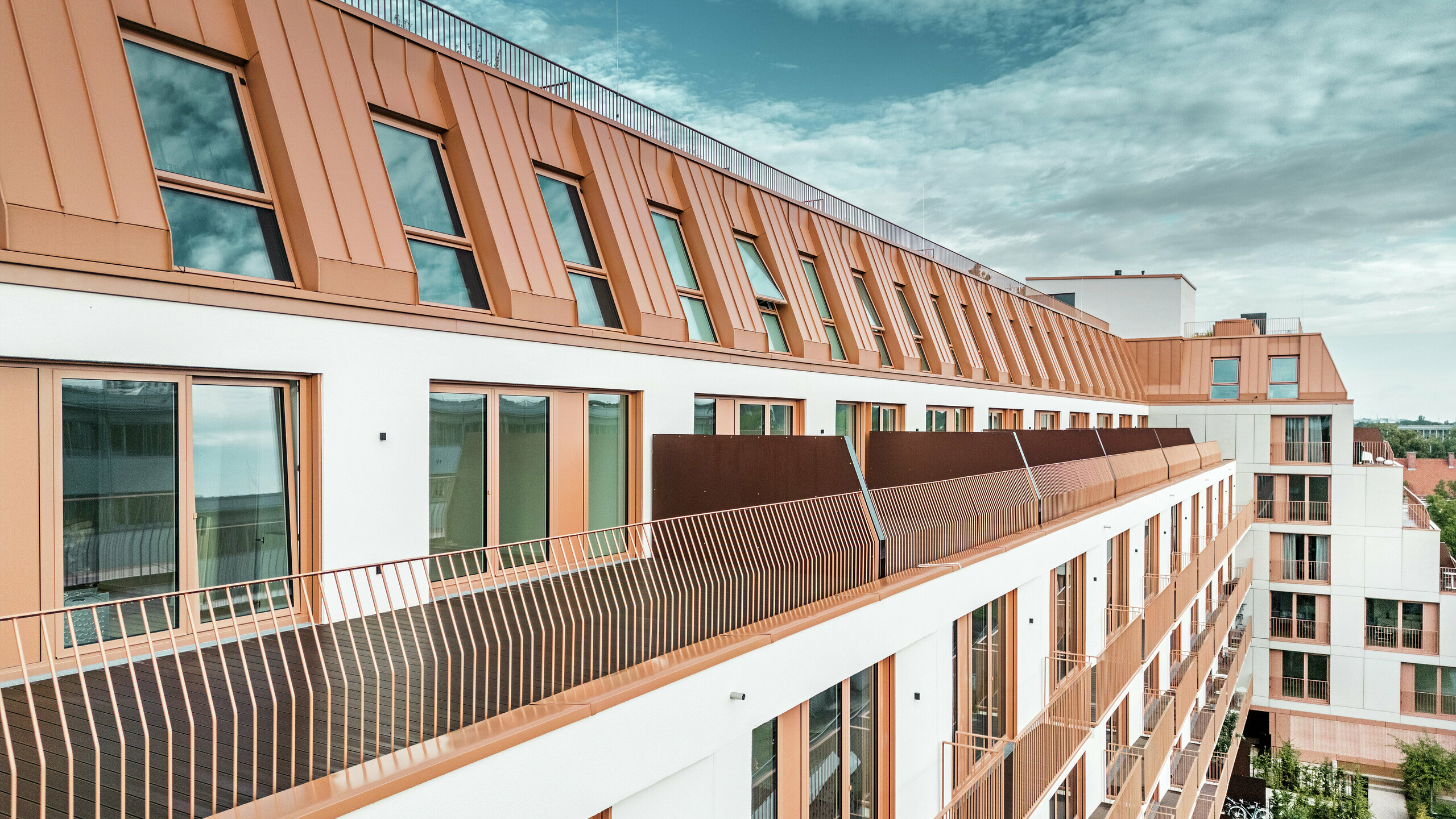 Immeuble résidentiel moderne Van B à Munich avec un paysage de toit saisissant en aluminium PREFALZ dans la couleur spéciale IGP Orange HDP. La couverture de toit durable et nécessitant peu d'entretien de PREFA garantit une unité esthétique et une grande résistance aux intempéries. La conception élégante de la façade avec ses cadres de fenêtres cuivrés-orange et ses généreux balcons souligne le concept architectural innovant. La solution idéale pour les projets de construction urbaine avec des exigences élevées en matière de design et de fonctionnalité.