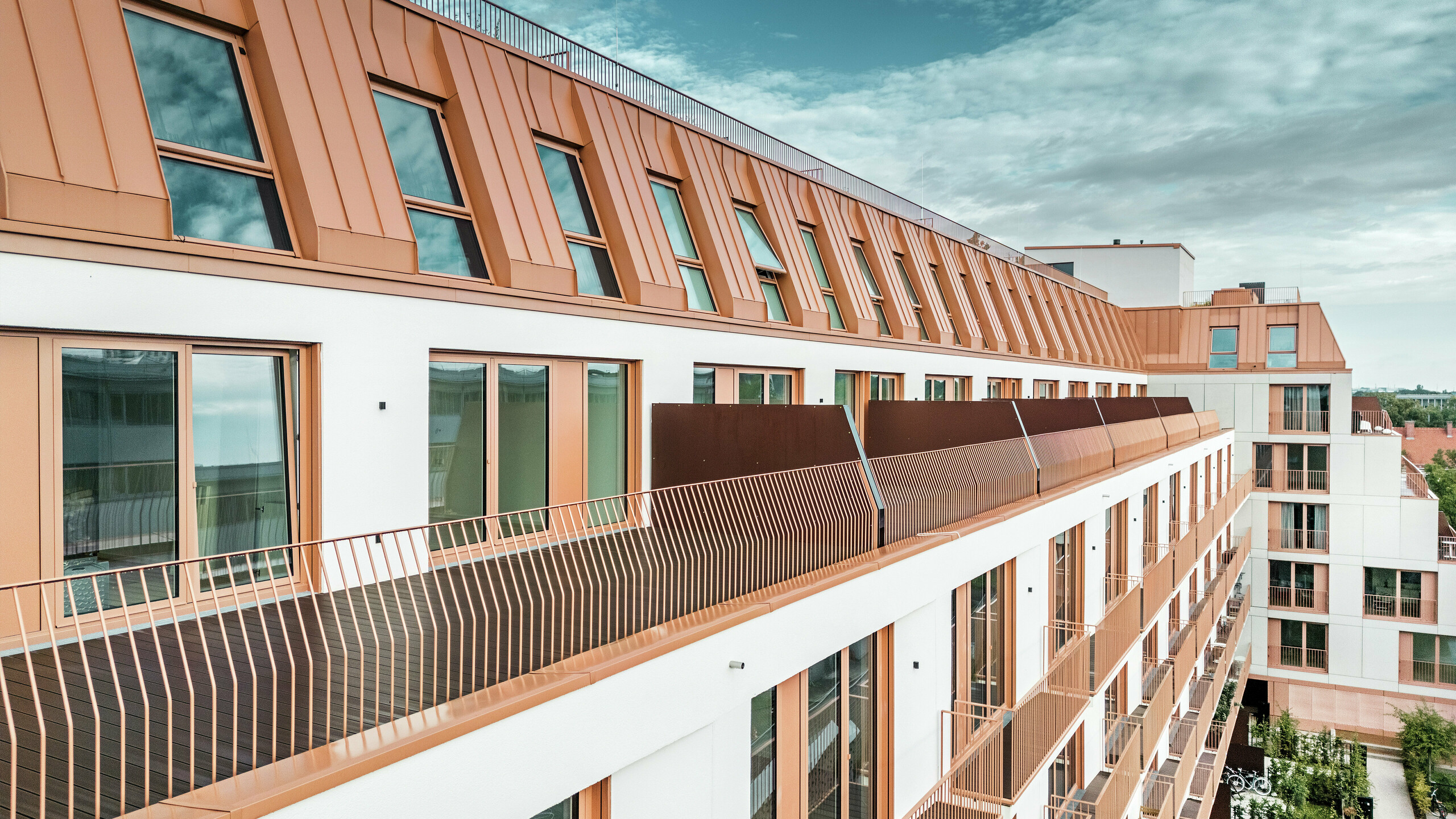 Immeuble résidentiel moderne Van B à Munich avec un paysage de toit saisissant en aluminium PREFALZ dans la couleur spéciale IGP Orange HDP. La couverture de toit durable et nécessitant peu d'entretien de PREFA garantit une unité esthétique et une grande résistance aux intempéries. La conception élégante de la façade avec ses cadres de fenêtres cuivrés-orange et ses généreux balcons souligne le concept architectural innovant. La solution idéale pour les projets de construction urbaine avec des exigences élevées en matière de design et de fonctionnalité.