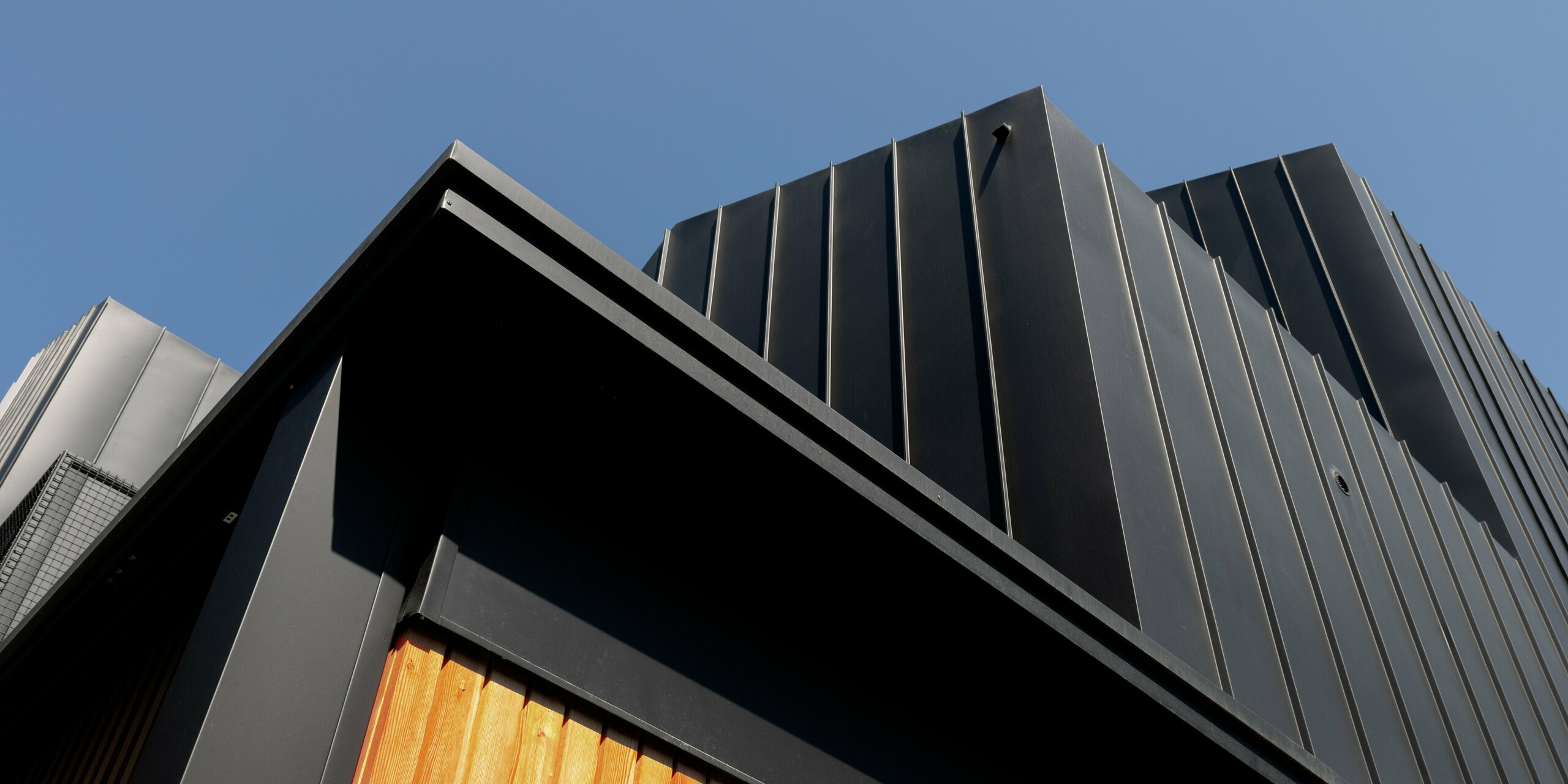 Gros plan sur l'architecture moderne de Cattolica : une façade en aluminium PREFALZ gris noir remarquable, associée à un bardage en bois chaleureux. La pureté des lignes et la qualité supérieure de la surface PREFA garantissent durabilité, protection contre la corrosion et un design élégant et intemporel sous un ciel d'un bleu éclatant.