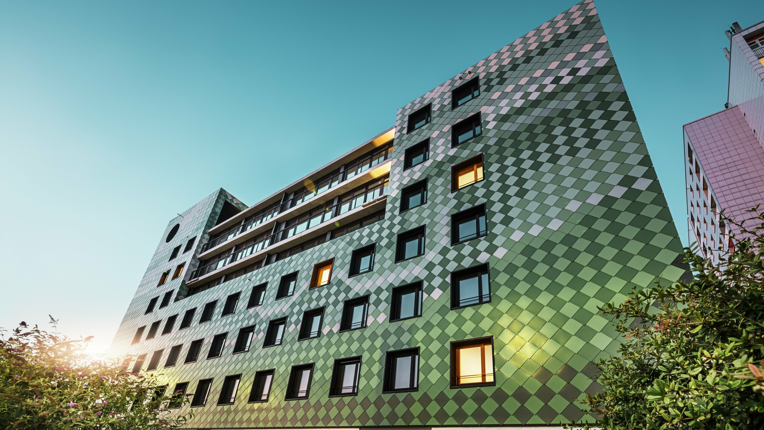 Bâtiment résidentiel et commercial moderne "Pick-Nick" à Dresde avec une façade PREFA frappante en losanges d'aluminium dans des couleurs spéciales vert olive, vert gris, vert réséda, vert opale, ivoire clair, blanc perle et bronze clair. Le traitement créatif de la façade rappelle une rizière et crée un jeu de couleurs vivant. L'architecture claire, les cadres de fenêtres noirs et la durabilité des matériaux PREFA façonnent le bâtiment. Le soleil bas se reflète sur la façade et les surfaces des fenêtres.