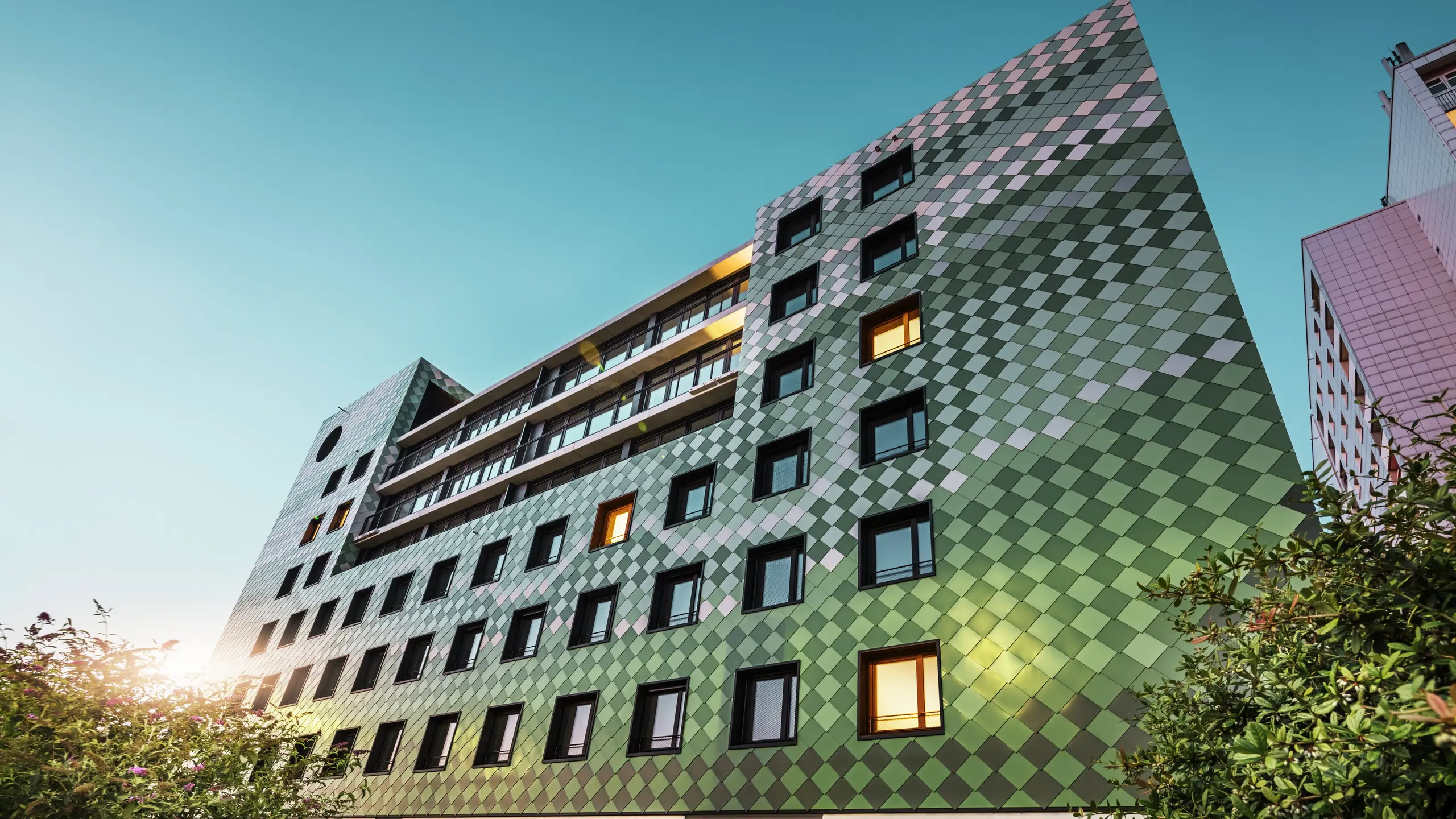 Bâtiment résidentiel et commercial moderne "Pick-Nick" à Dresde avec une façade PREFA frappante en losanges d'aluminium dans des couleurs spéciales vert olive, vert gris, vert réséda, vert opale, ivoire clair, blanc perle et bronze clair. Le traitement créatif de la façade rappelle une rizière et crée un jeu de couleurs vivant. L'architecture claire, les cadres de fenêtres noirs et la durabilité des matériaux PREFA façonnent le bâtiment. Le soleil bas se reflète sur la façade et les surfaces des fenêtres.