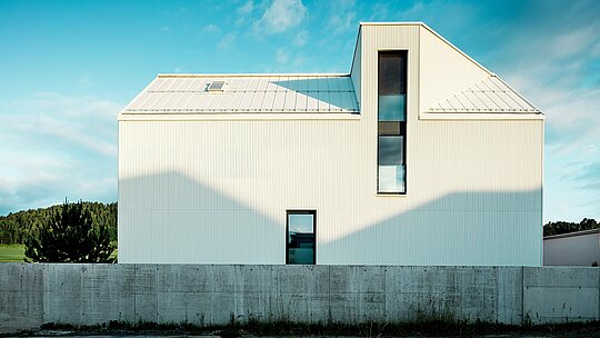 Cette maison individuelle moderne de Freistadt a été photographiée de profil afin de mettre en valeur le design remarquable de sa façade et de sa toiture. Le bardage en aluminium blanc pur PREFALZ, associé à la toiture à forte pente et aux fenêtres étroites, confère au bâtiment une allure à la fois moderne et intemporelle. Les rainures verticales de la façade lui donnent un aspect texturé et dynamique. Le jeu d'ombre et de lumière crée des effets d'ombre intéressants et souligne la précision des lignes. Cette architecture épurée et minimaliste est sublimée par la façade PREFALZ, durable et résistante aux intempéries, reconnue pour sa longévité et sa facilité d'entretien.