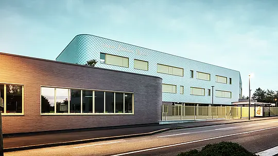 Vue du soir du collège Léontine Dolivet à Cesson-Sévigné. Au premier plan, une rue bien délimitée longe un immeuble bas aux larges baies vitrées. Derrière, se dresse le bâtiment principal, dont la façade incurvée en aluminium, composée de losanges PREFA argent métallisé, scintille sous la douce lumière. Plusieurs fenêtres rectangulaires et une clôture claire structurent la composition. La scène est éclairée par les lampadaires, et le ciel d'un bleu frais offre un spectacle moderne et élégant.