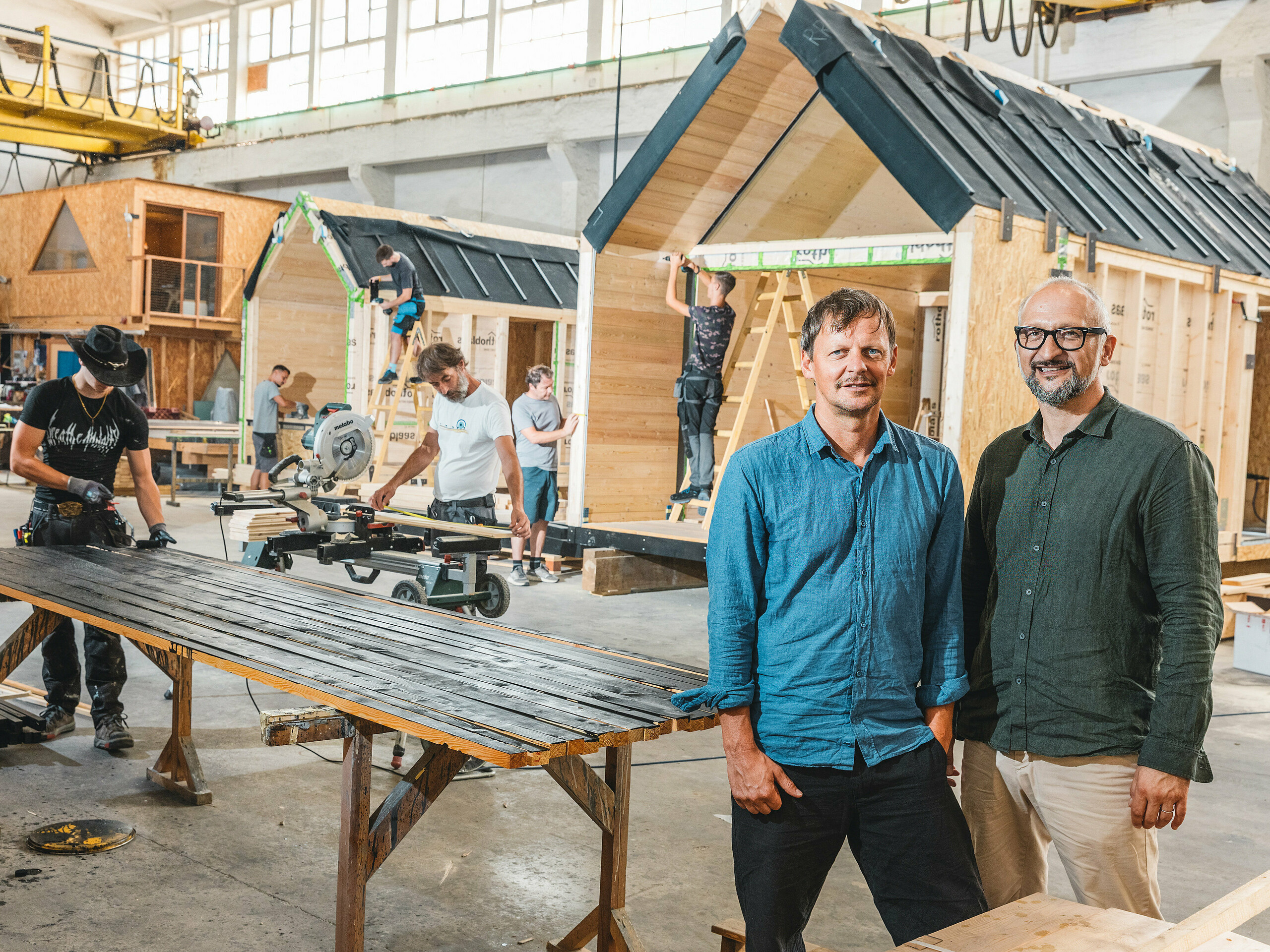 Dans un atelier de production, les maisons miniatures minimalistes de Lushna sont en cours d'assemblage. L'entreprise, qui jouit d'une renommée internationale, est spécialisée dans l'architecture miniature pour les complexes hôteliers en pleine nature. Au premier plan, on aperçoit les deux fondateurs, l'architecte Peter Ličen et le spécialiste marketing Jaka Ažman. À l'arrière-plan, des maisons à ossature en A sont assemblées. Le contraste saisissant entre le bois et l'aluminium foncé confère à l'architecture un aspect visuel remarquable, comme en témoigne le Jardin secret de carpes koï du lac Natterer.