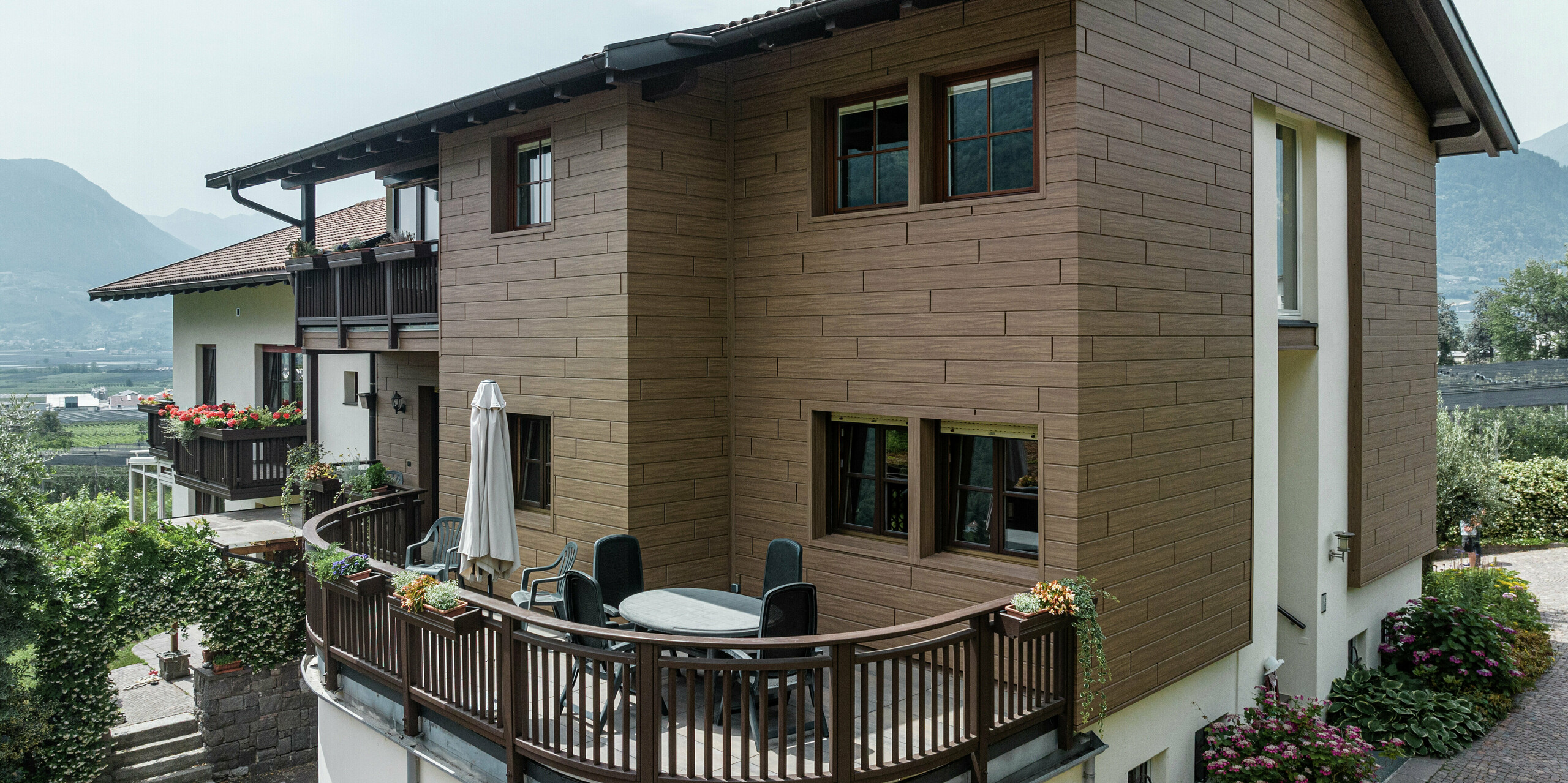 Appartements de vacances rénovés Prechtlhof à Merano, Tyrol du Sud, avec sIDING PREFA en noyer foncé. La façade en aluminium sans entretien à l'aspect bois allie esthétique moderne et charme traditionnel et s'intègre harmonieusement dans l'environnement alpin. Le balcon circulaire au premier plan vous invite à vous attarder dans le magnifique paysage du Tyrol du Sud.