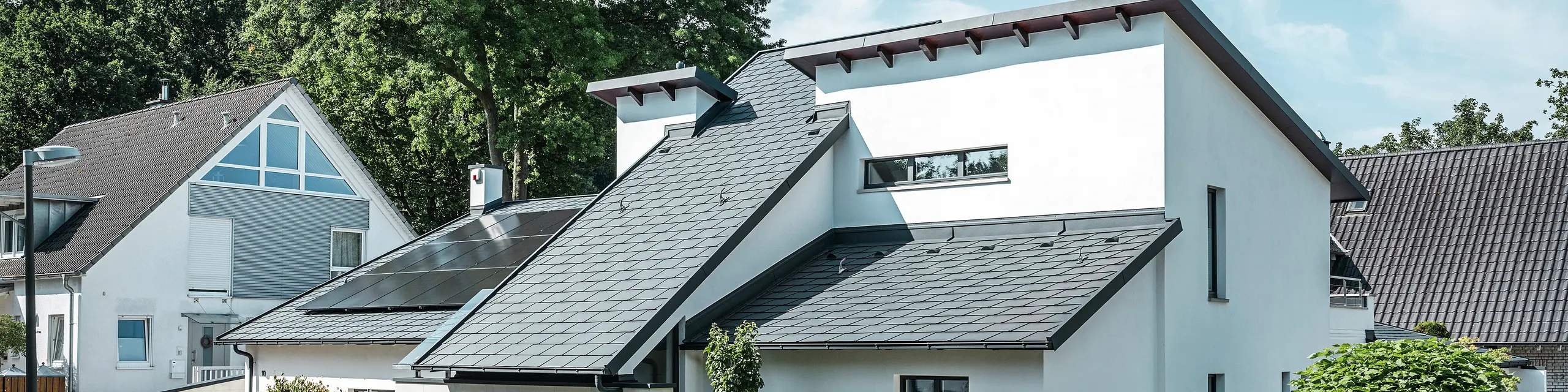Maison individuelle moderne avec façade en crépi blanc et toit en pente PREFA en aluminium foncé. La toiture est équipée de modules photovoltaïques intégrés et de gouttières en aluminium anthracite. Le bâtiment est situé dans un lotissement bien entretenu avec des jardins fleuris, une allée pavée et des maisons individuelles environnantes.