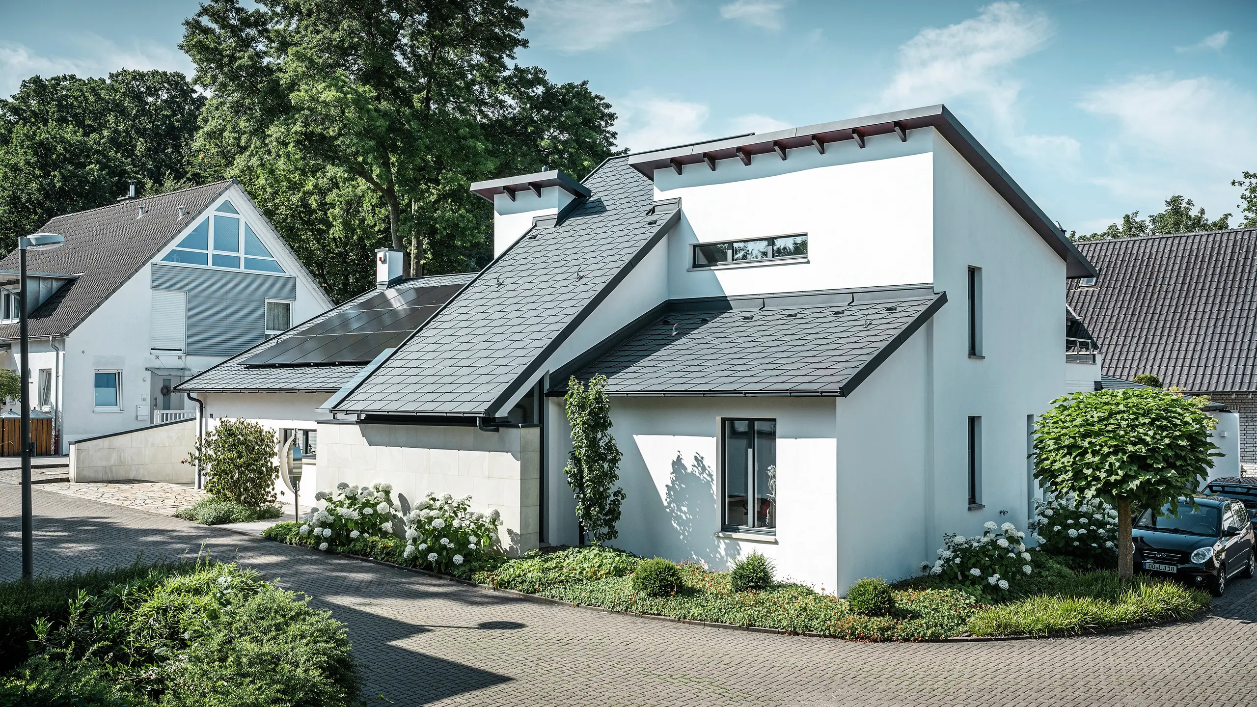Maison individuelle moderne avec façade en crépi blanc et toit en pente PREFA en aluminium foncé. La toiture est équipée de modules photovoltaïques intégrés et de gouttières en aluminium anthracite. Le bâtiment est situé dans un lotissement bien entretenu avec des jardins fleuris, une allée pavée et des maisons individuelles environnantes.