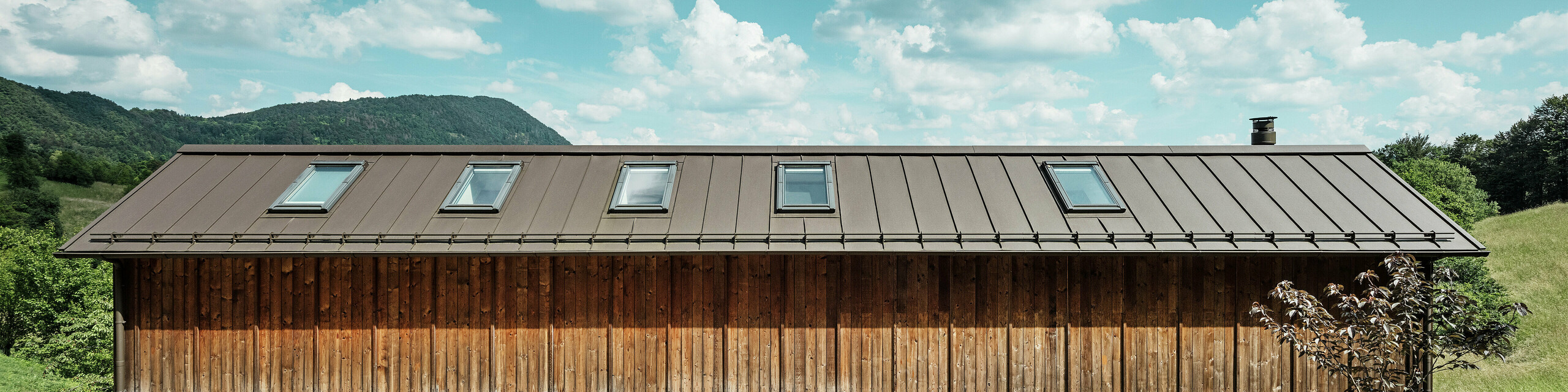 Cette maison moderne construite en pente séduit par sa façade en bois minimaliste et son toit en aluminium PREFALZ durable de couleur P.10 brun. La couverture à joint debout convainc par sa grande résistance aux intempéries et sa longévité, ainsi que par son harmonie avec l’environnement naturel. Des fenêtres de toit intégrées et des lignes épurées soulignent l’architecture intemporelle. Sur la photo, on distingue l’entrée de cette petite maison individuelle.