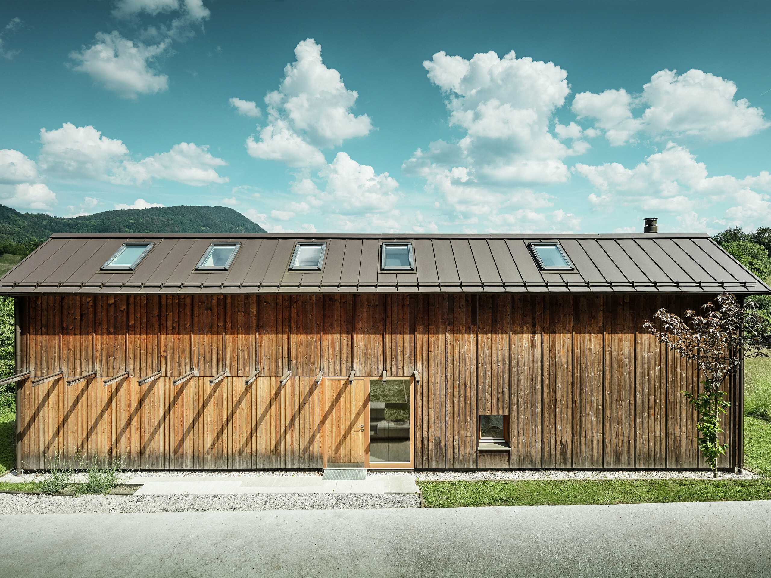 Cette maison moderne construite en pente séduit par sa façade en bois minimaliste et son toit en aluminium PREFALZ durable de couleur P.10 brun. La couverture à joint debout convainc par sa grande résistance aux intempéries et sa longévité, ainsi que par son harmonie avec l’environnement naturel. Des fenêtres de toit intégrées et des lignes épurées soulignent l’architecture intemporelle. Sur la photo, on distingue l’entrée de cette petite maison individuelle.