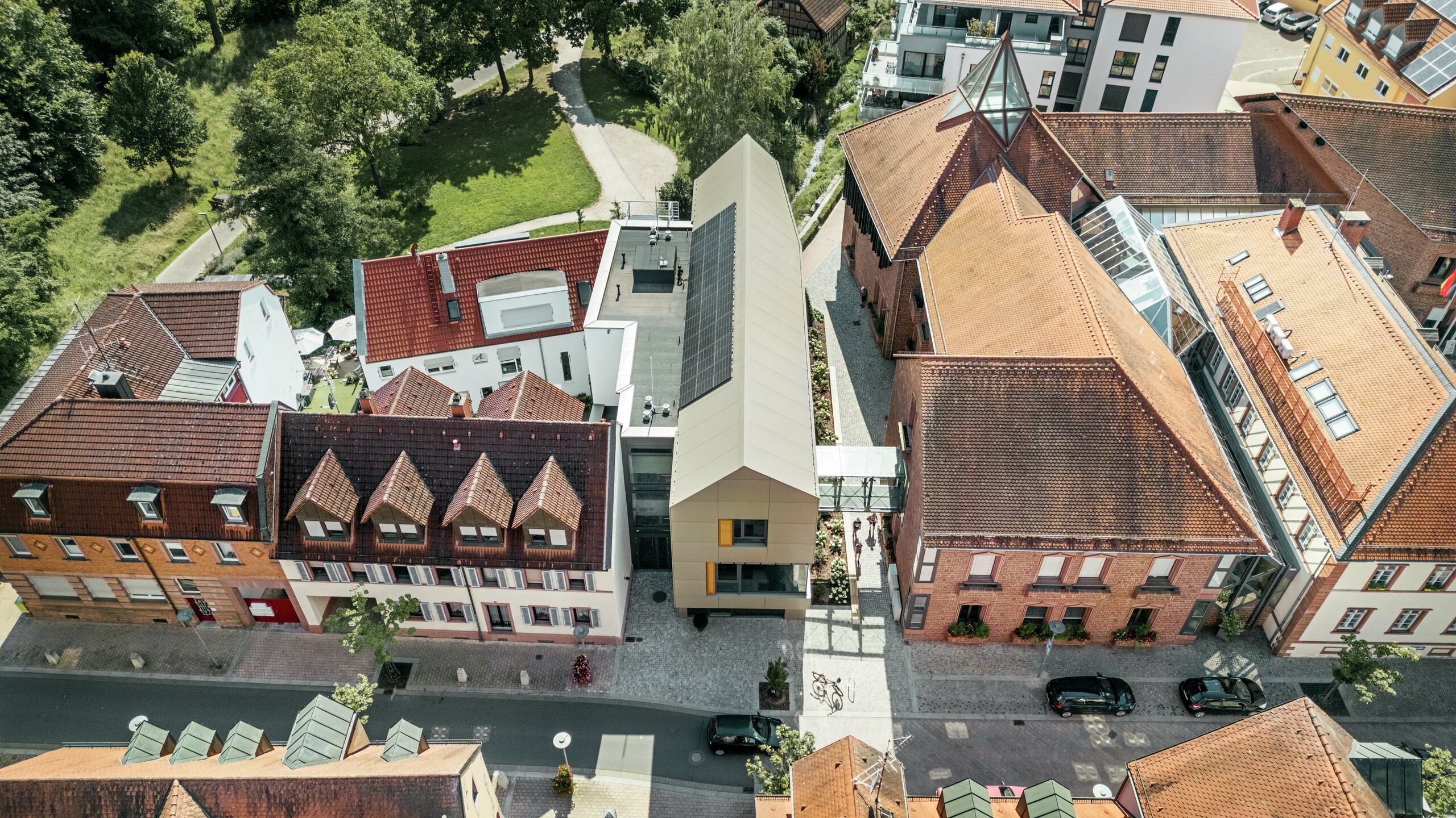 La mairie d'Alzenau, en Allemagne, est située au centre d'un paysage urbain historique, entourée de bâtiments traditionnels aux toits de tuiles rouges. La mairie moderne contraste avec ces bâtiments, sa façade étant revêtue de panneaux composites en aluminium PREFA dans un élégant ton bronze. L'architecture innovante se démarque clairement des bâtiments classiques voisins tout en s'intégrant harmonieusement dans le paysage urbain.