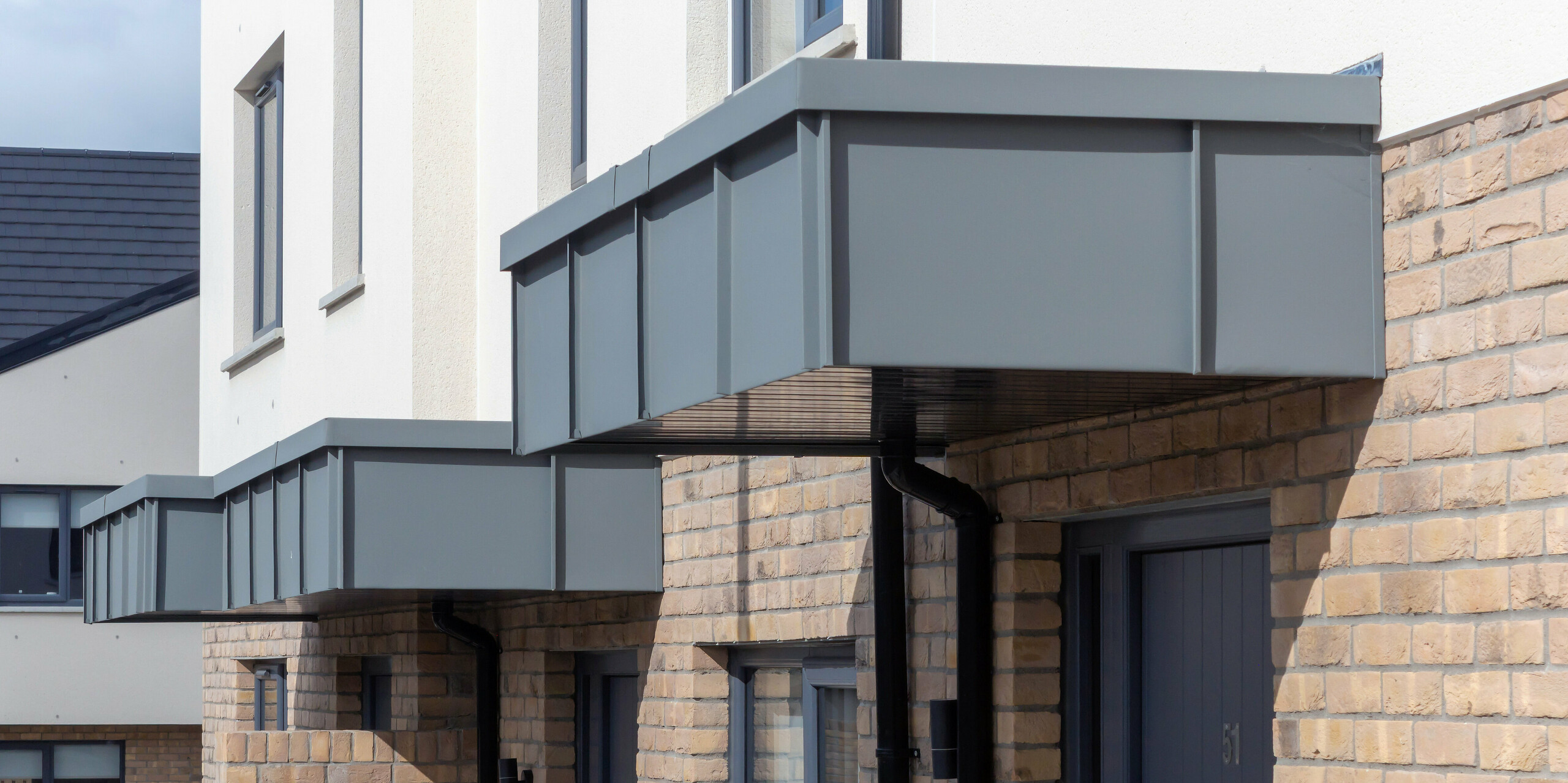 L'image présente les auvents d'entrée remarquables des maisons mitoyennes de Churchfields, entièrement revêtus de PREFALZ couleur P.10 gris souris. Les lignes épurées et droites des surfaces en aluminium créent un contraste moderne avec la chaleur de la brique et confèrent à l'ensemble une apparence uniforme et haut de gamme. La surface robuste de PREFA est résistante aux intempéries, inaltérable et ne nécessite pratiquement aucun entretien – idéale pour les bâtiments résidentiels durables. Le gros plan met en valeur la précision de la pose et l'intégration harmonieuse du revêtement en aluminium à l'architecture.