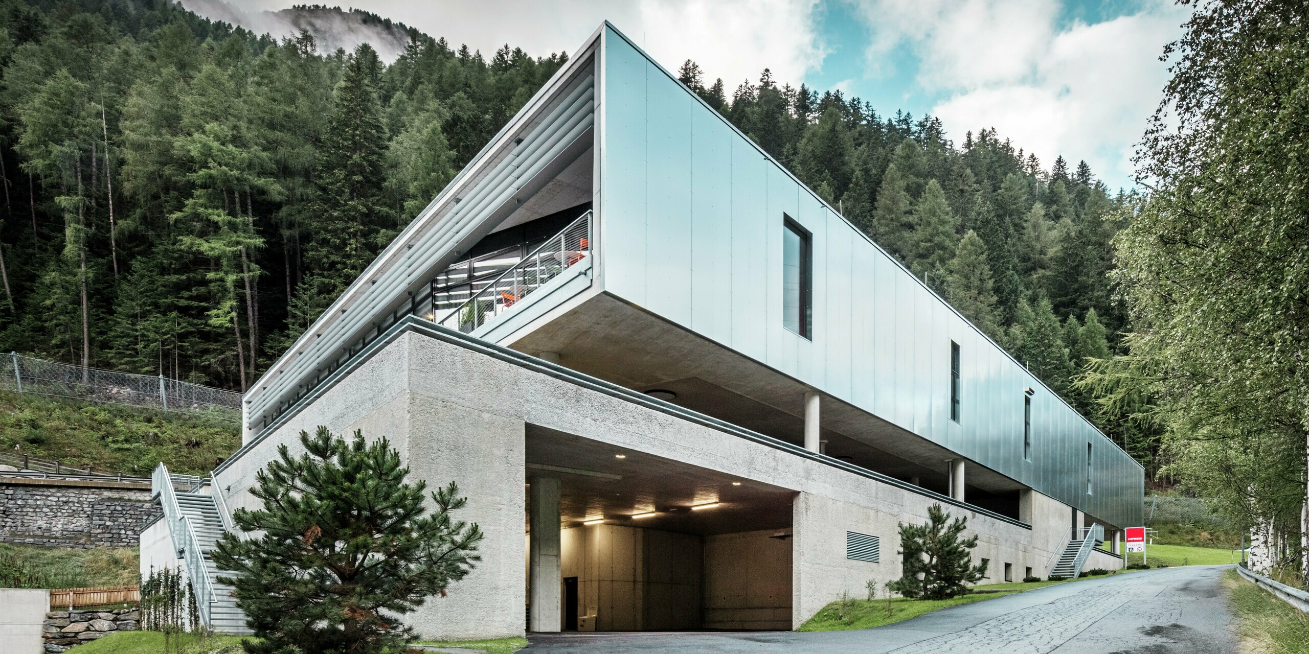 Vue arrière du magasin MPREIS à Sölden, au Tyrol, avec des panneaux composites en aluminium PREFA réfléchissants dans la couleur spéciale Reflective Natural. L'architecture moderne s'intègre dans le paysage alpin boisé et allie fonctionnalité et esthétique. Le parking et la route d'accès constituent une solution pratique pour construire sur un terrain en pente.