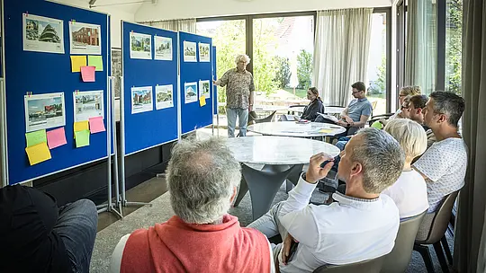 PREFARENZEN 2027 : Dialogue d’experts dans une salle de séminaire moderne et lumineuse : Matthias Finkentey, modérateur, se tient devant des panneaux d’affichage bleus présentant des projets architecturaux intégrant les solutions aluminium PREFA. Le jury, assis autour de tables rondes, discute et prend des notes. De larges fenêtres laissent entrer la lumière naturelle, tandis qu’à l’extérieur, un jardin verdoyant s’offre à la vue. Cette scène illustre le lien entre réflexion architecturale créative, planification fonctionnelle et évaluation du design. Les produits aluminium PREFA offrent des toitures et des façades durables et esthétiques, incarnant l’innovation, la qualité et l’inspiration dans les projets architecturaux.