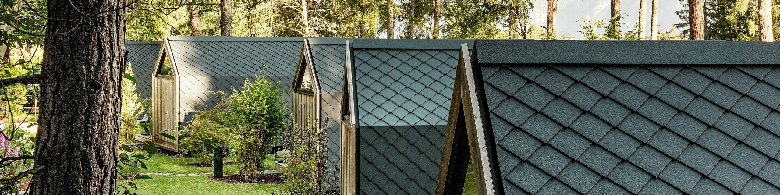 Plusieurs mini-maisons minimalistes du Secret Koi Garden s'harmonisent le long d'un axe verdoyant bordant le lac Natterer. Toits et façades sont entièrement revêtus de losanges en aluminium PREFA P.10 anthracite, créant une surface uniforme évoquant des écailles de poisson. Entourées d'arbres et d'arbustes, ces maisons allient une architecture précise, la tranquillité de la nature et un design glamping haut de gamme, grâce aux produits PREFA en aluminium résistants aux intempéries.