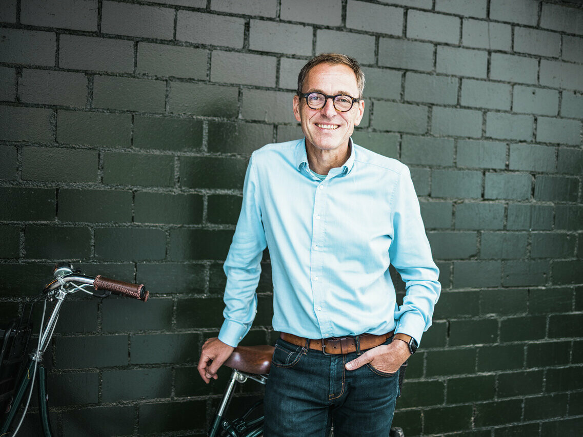 Portrait de l'architecte Jochen Weissenrieder, adossé à son vélo, avec un mur en brique noir en arrière-plan.