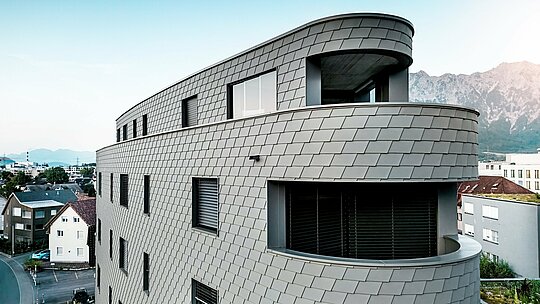Vue oblique détaillée de la Bernerhaus à Buchs, révélant un angle légèrement arrondi et des balcons continus. La façade, revêtue de bardeaux PREFA gris souris P.10, épouse la structure triangulaire comme une peau d'aluminium finement travaillée, créant un motif vibrant évoquant des écailles. L'homogénéité de la surface souligne les courbes harmonieuses et le jeu d'ombre et de lumière. Grâce à la légèreté, la résistance aux intempéries et la durabilité de l'aluminium PREFA, l'enveloppe du bâtiment, nécessitant peu d'entretien, offre un impact visuel saisissant sur le fond alpin.