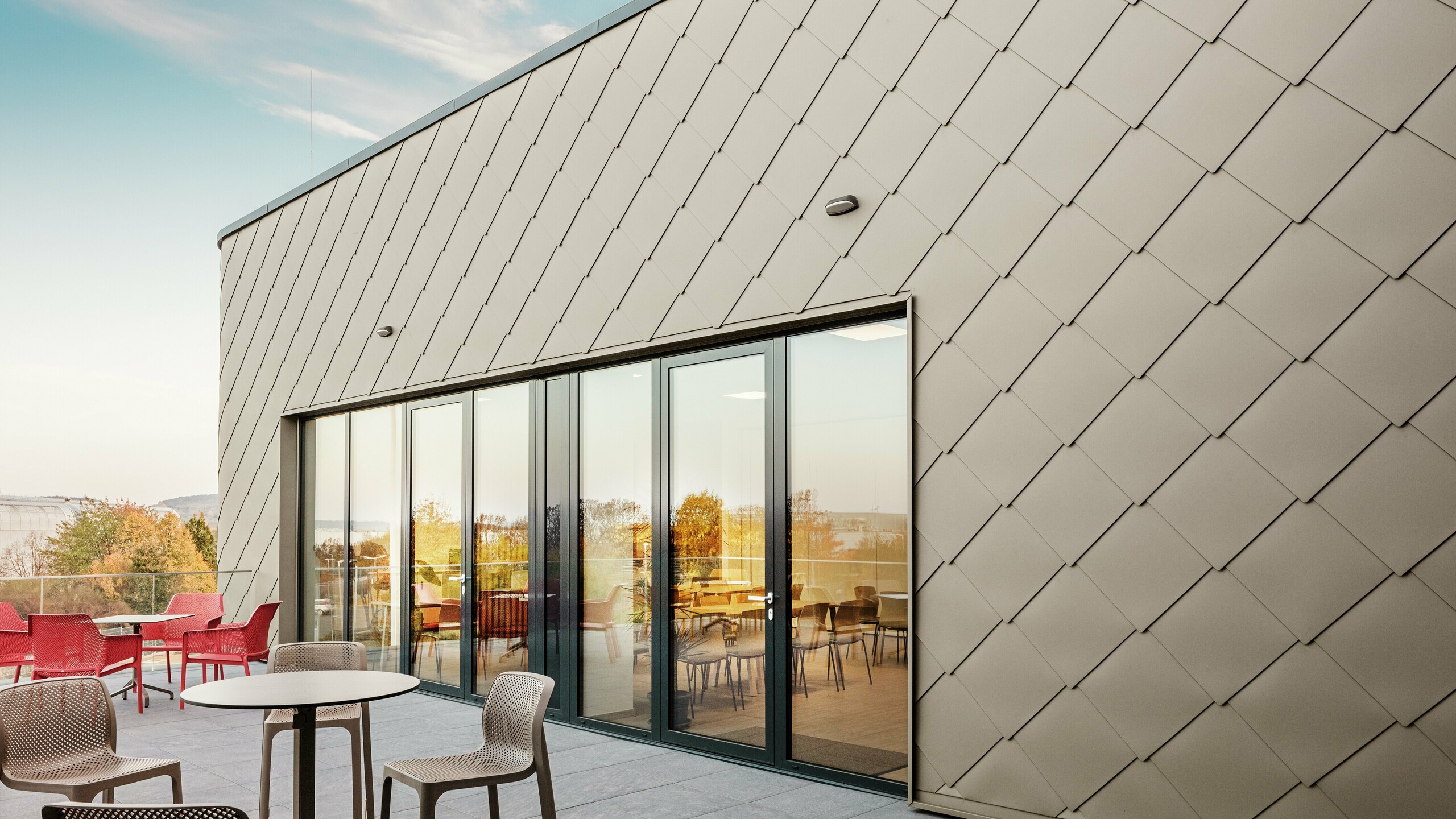 La façade du nouveau site PREFA à Törökbálint impressionne par les élégants losanges de façade PREFA 44 × 44 en bronze. La façade en aluminium posée avec précision crée une apparence moderne et dynamique tout en offrant une protection maximale contre les éléments. La généreuse façade vitrée relie les espaces intérieurs et extérieurs et crée une atmosphère lumineuse et accueillante sur la terrasse spacieuse.
