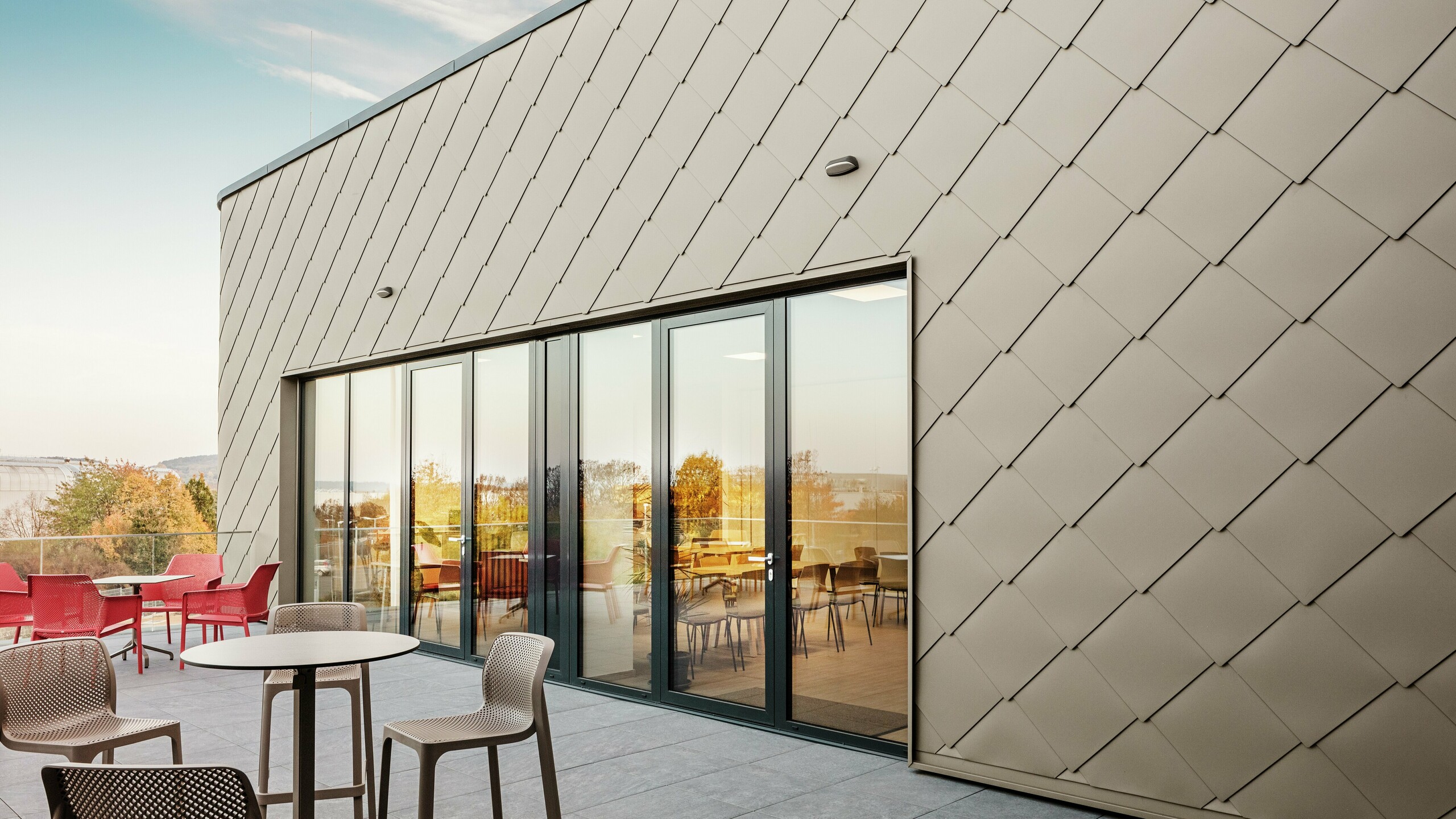 La façade du nouveau site PREFA à Törökbálint impressionne par les élégants losanges de façade PREFA 44 × 44 en bronze. La façade en aluminium posée avec précision crée une apparence moderne et dynamique tout en offrant une protection maximale contre les éléments. La généreuse façade vitrée relie les espaces intérieurs et extérieurs et crée une atmosphère lumineuse et accueillante sur la terrasse spacieuse.
