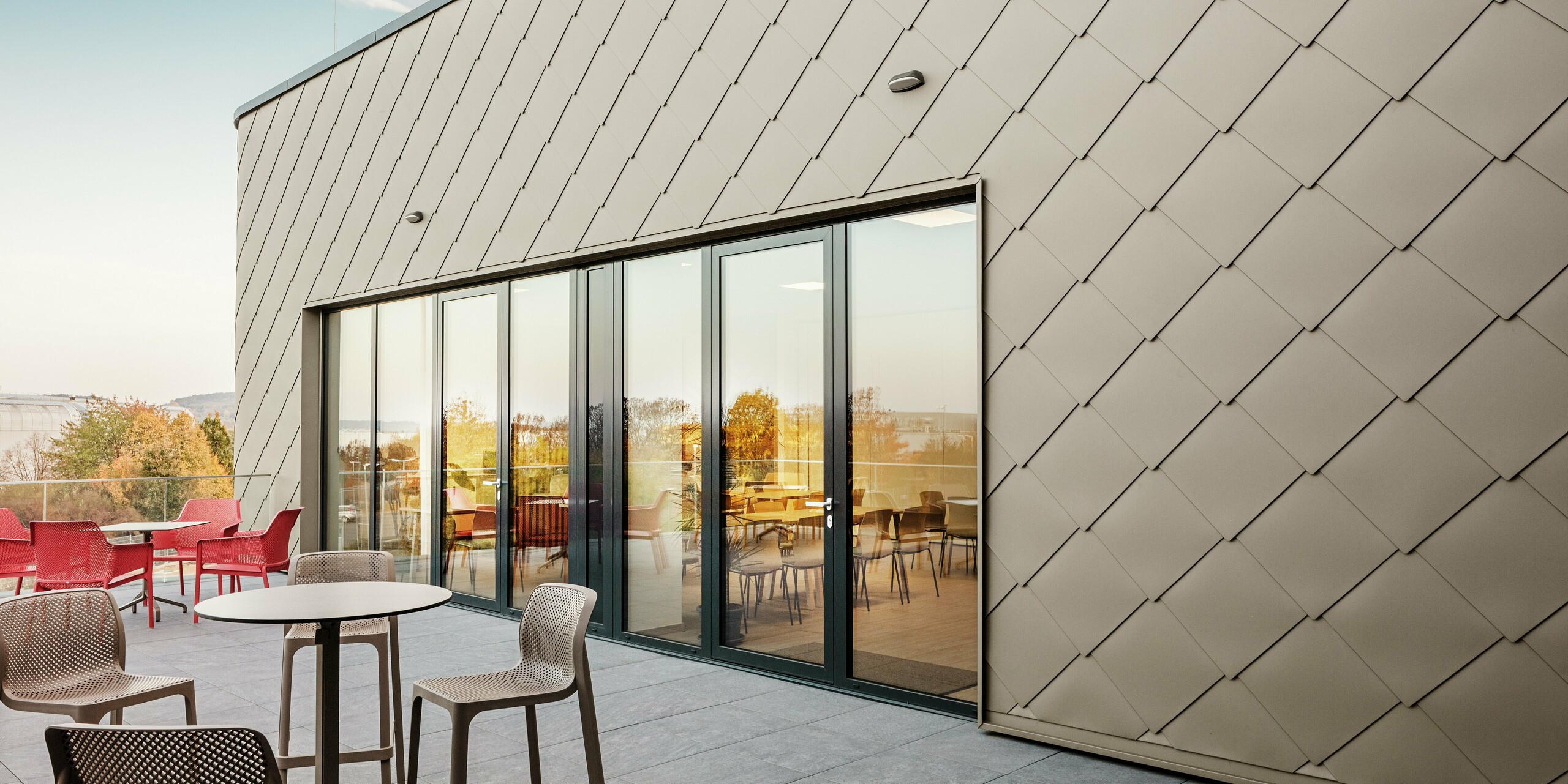 La façade du nouveau site PREFA à Törökbálint impressionne par les élégants losanges de façade PREFA 44 × 44 en bronze. La façade en aluminium posée avec précision crée une apparence moderne et dynamique tout en offrant une protection maximale contre les éléments. La généreuse façade vitrée relie les espaces intérieurs et extérieurs et crée une atmosphère lumineuse et accueillante sur la terrasse spacieuse.