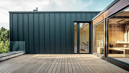 La photographie révèle la vaste terrasse sur le toit d'une villa multigénérationnelle à Budapest, dont la façade est revêtue d'aluminium PREFALZ couleur P.10 anthracite. Les joints debout, précis et verticaux, confèrent à la façade une esthétique épurée et moderne, en parfaite harmonie avec le bois chaleureux du sauna adjacent. La lumière naturelle accentue l'élégant contraste des matériaux et met en valeur la finition PREFA, à la fois durable et de haute qualité. La généreuse terrasse offre une vue imprenable sur la verdure environnante et souligne le caractère moderne et axé sur le confort de la villa.