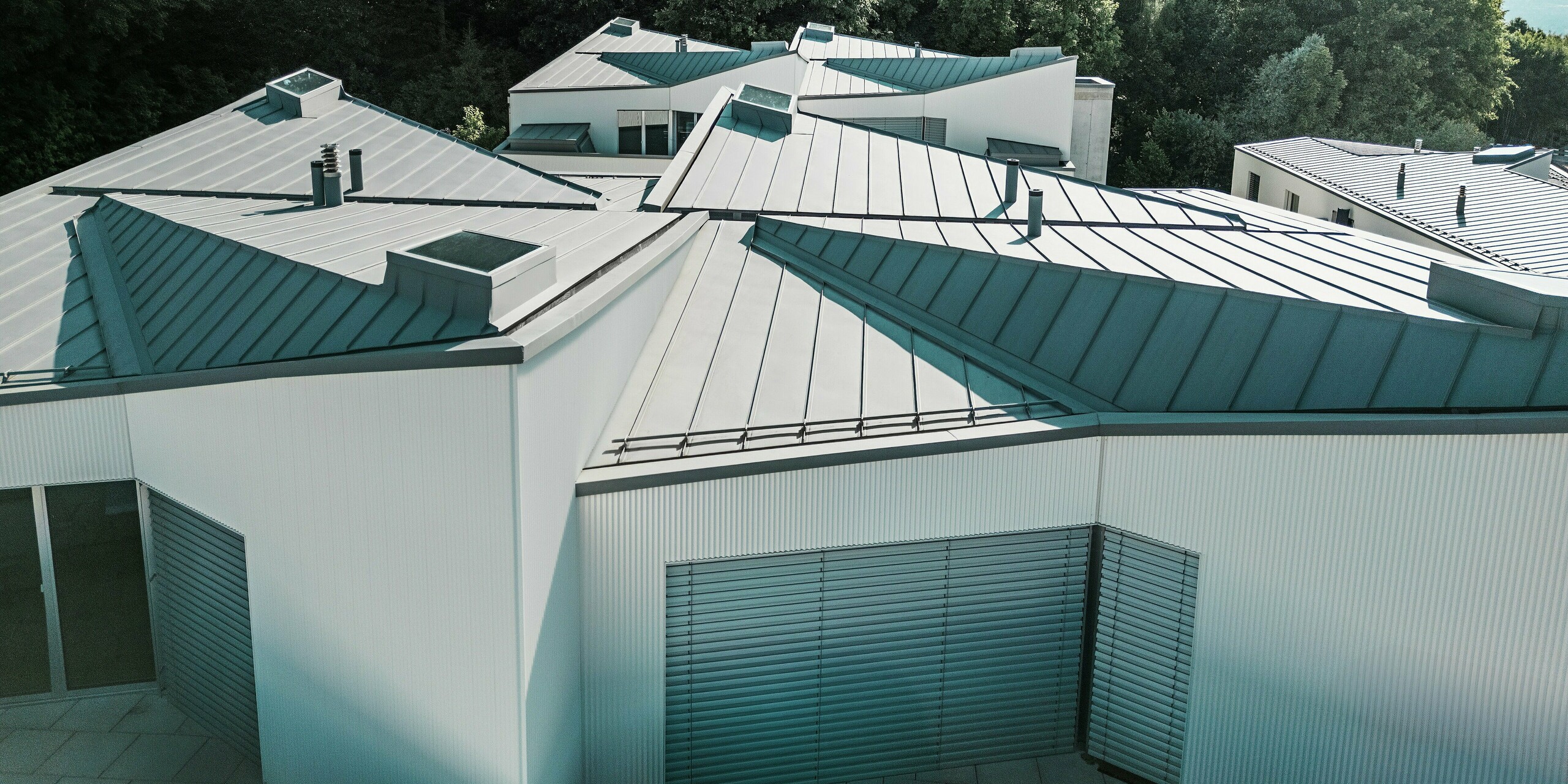 Vue panoramique des toitures PREFALZ exceptionnellement plissées, d'un gris patiné, surplombant un ensemble de mini-maisons modernes. Les surfaces à joints debout, réalisées avec précision, créent un jeu d'ombre et de lumière saisissant. L'aluminium PREFA, d'une grande durabilité, offre une protection fiable contre le vent et les intempéries et permet des conceptions de toiture architecturalement sophistiquées, même dans les espaces les plus réduits.