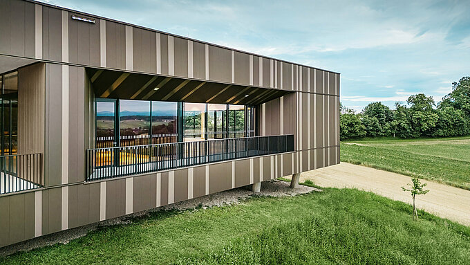 Vue latérale du centre événementiel ZIB Nikolai à St. Veit, avec une façade moderne en aluminium dans des tons verticaux de marron et de gris beige. La grande loggia aux fenêtres et garde-corps noirs s'ouvre sur le paysage verdoyant environnant. En arrière-plan, de vastes champs et une bande de forêt sous un ciel légèrement nuageux.
