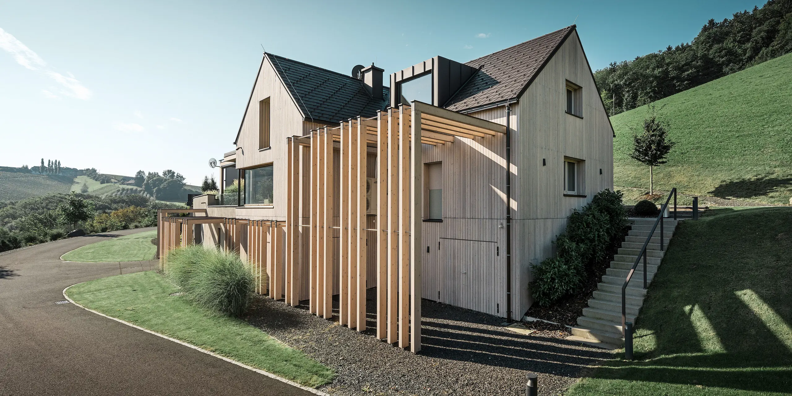 Grande maison individuelle avec toit en pente et lucarne, située à flanc de colline. La toiture est recouverte de bardeaux en aluminium brun PREFA et la façade est revêtue de planches verticales en bois.