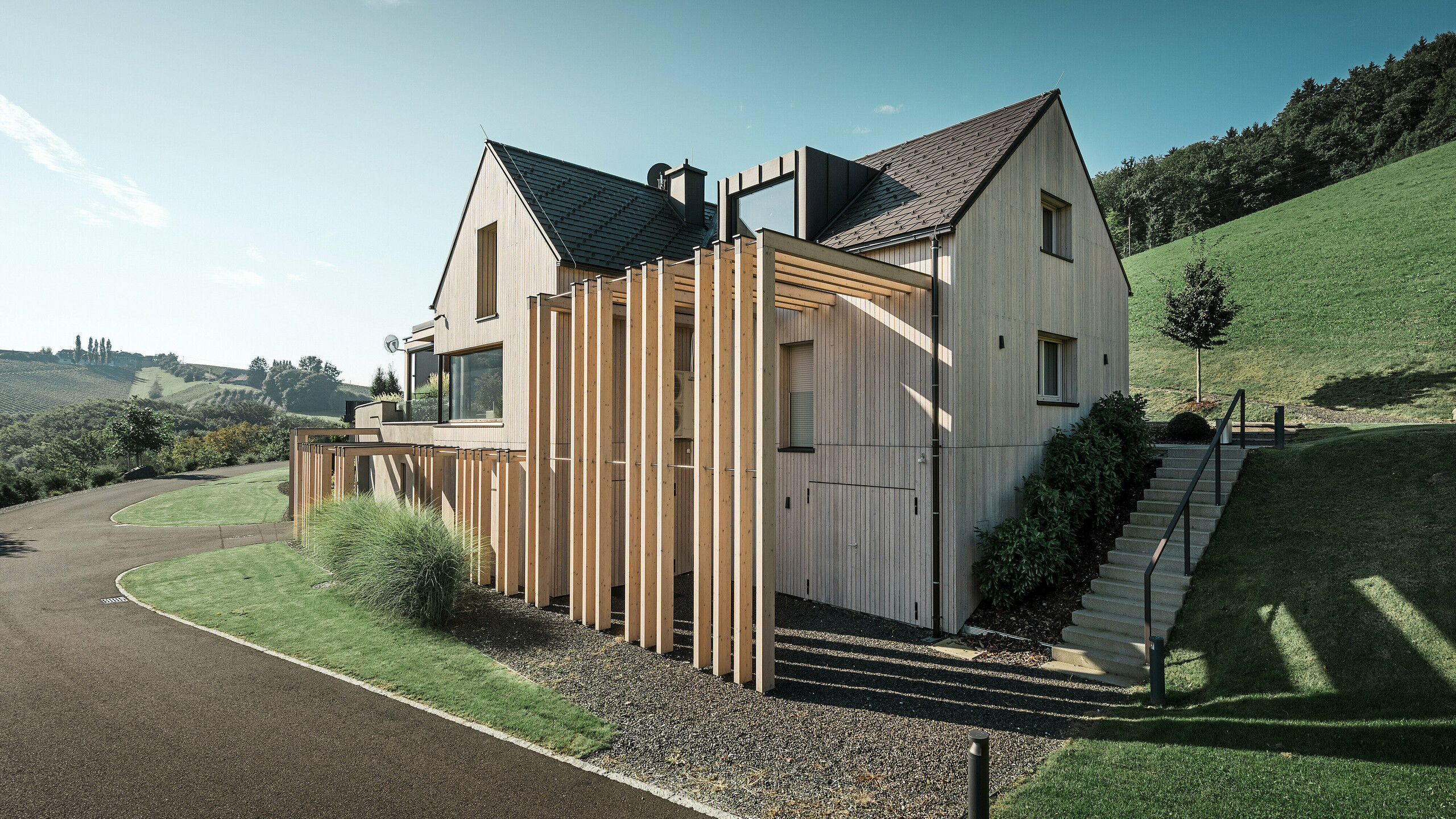Grande maison individuelle avec toit en pente et lucarne, située à flanc de colline. La toiture est recouverte de bardeaux en aluminium brun PREFA et la façade est revêtue de planches verticales en bois.