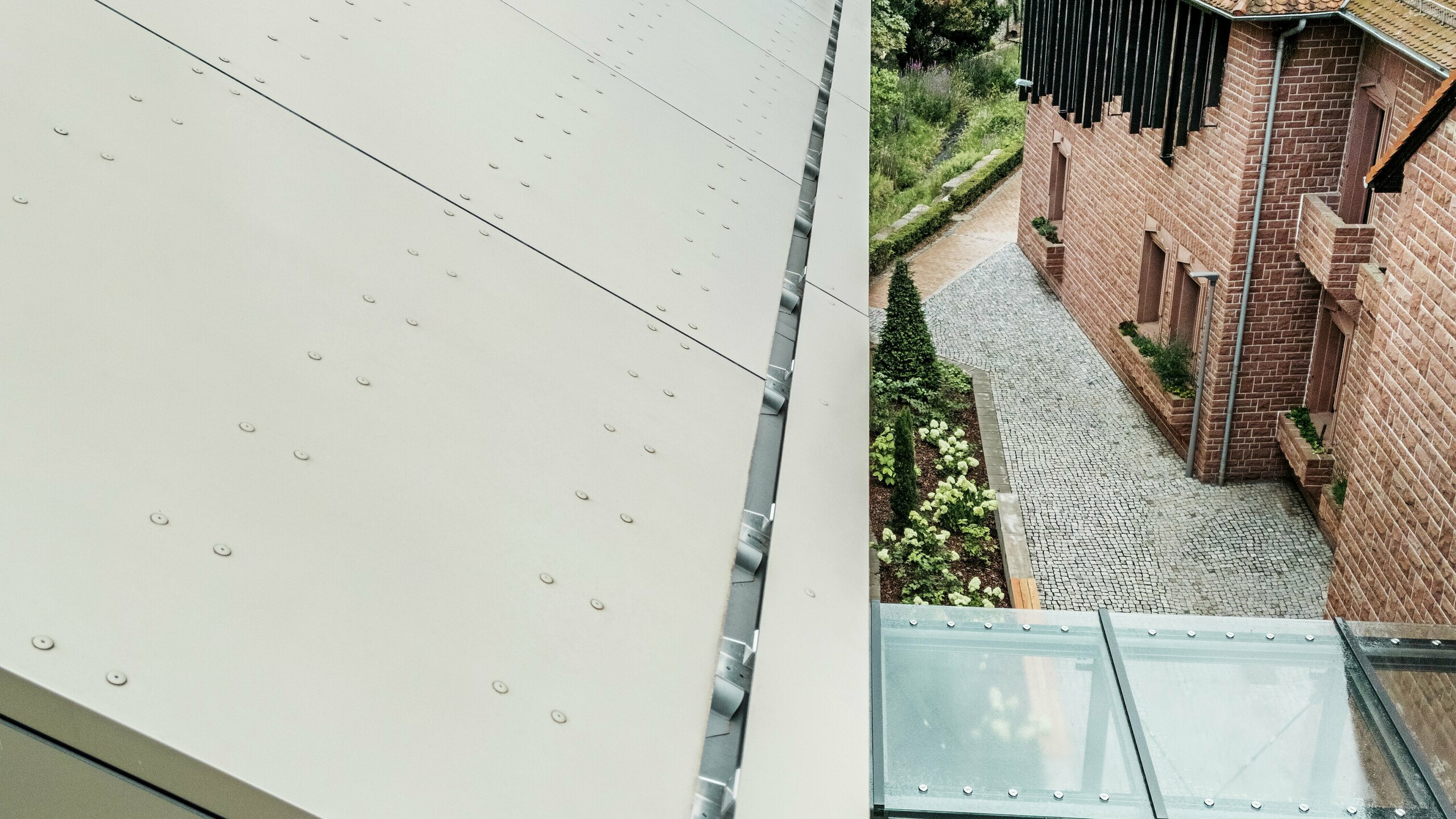 Détail de la toiture de la mairie d'Alzenau, recouverte de panneaux composites en aluminium PREFA dans une teinte bronze douce. Les panneaux sont posés avec précision et dotés d'éléments de fixation apparents, ce qui confère à la façade un aspect moderne et industriel. Au premier plan, on aperçoit une passerelle vitrée qui relie la mairie à un bâtiment voisin.