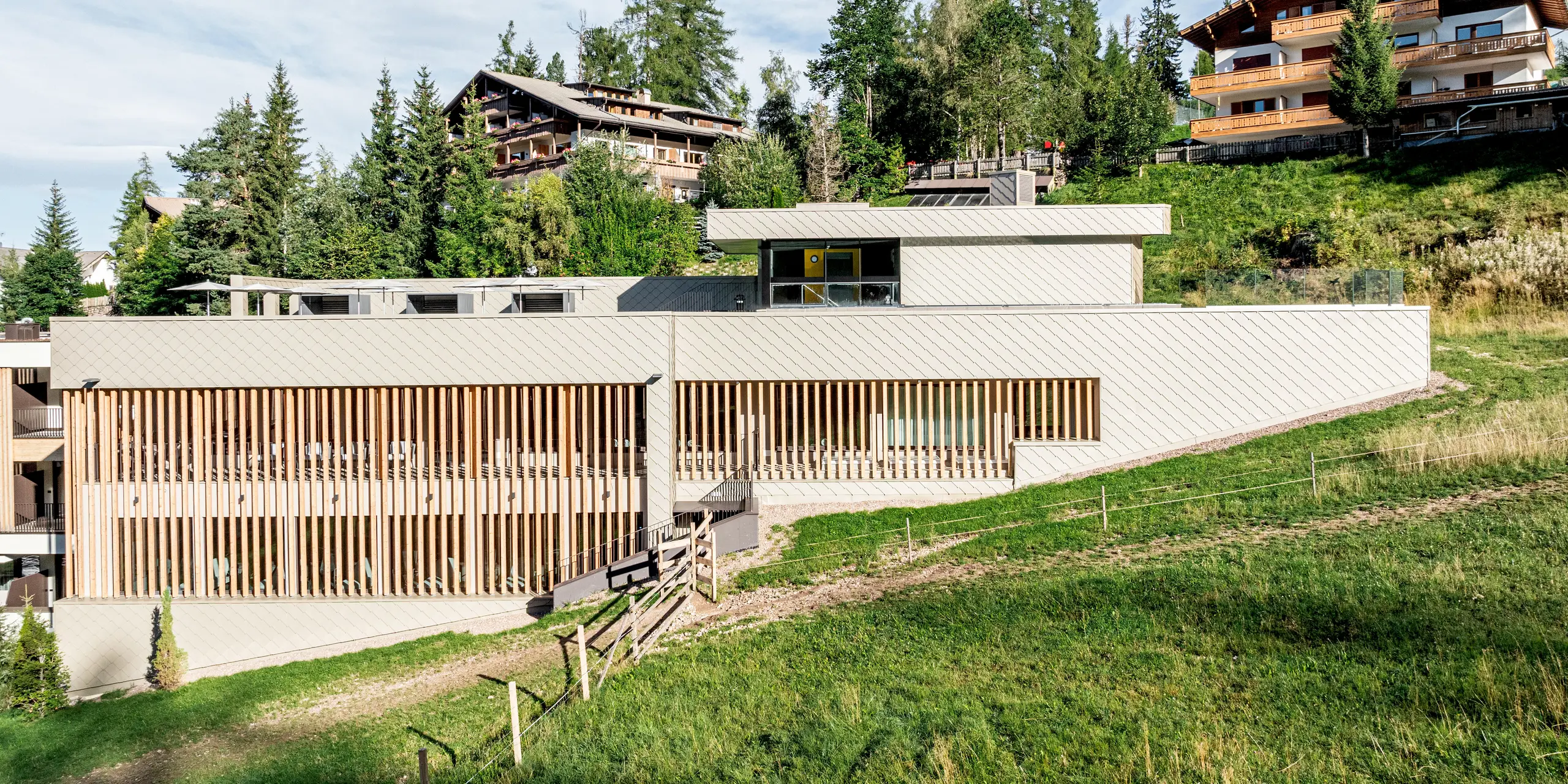 Vue latérale du Sporthotel Obereggen, situé dans le Tyrol du Sud, niché sur un doux versant alpin au milieu des prairies, des arbres et des bâtiments de montagne environnants. La structure allongée est caractérisée par une façade en aluminium PREFA composée de losanges de façade 29x29 couleur bronze, conférant à l'architecture des lignes épurées et une texture dynamique. La teinte chaude du bronze contraste harmonieusement avec les lattes de bois et le paysage naturel. Durables, résistants aux intempéries et nécessitant peu d'entretien, les produits en aluminium PREFA sont parfaitement adaptés à une architecture raffinée en régions alpines.
