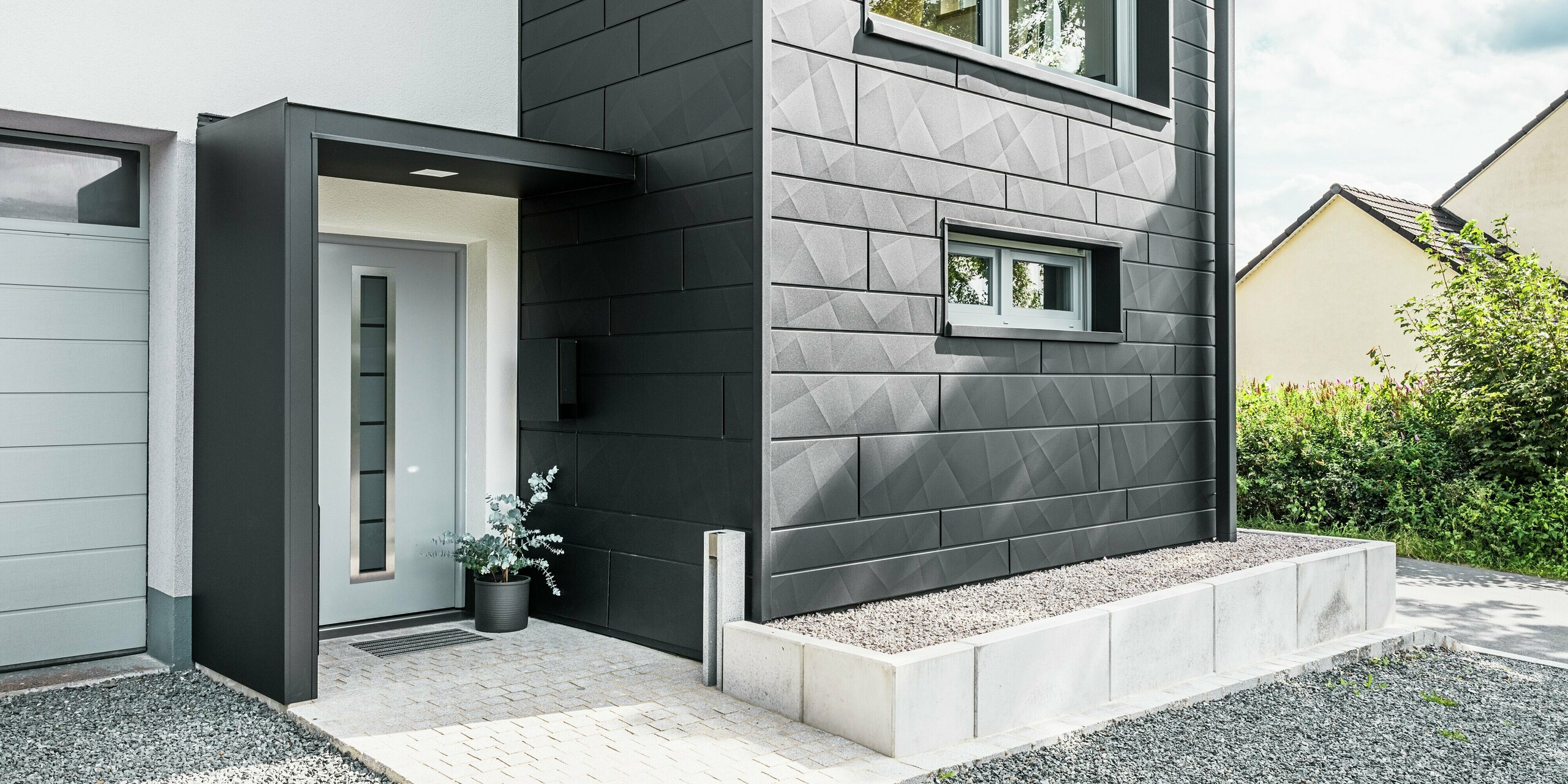 L'élégante façade Siding.X PREFA en P.10 noir domine l'entrée de la maison individuelle modernisée de Teuschnitz. La façade en aluminium de haute qualité confère au bâtiment un caractère moderne et intemporel. Les panneaux de façade clairement structurés créent un design géométrique saisissant, tandis que les panneaux composites en aluminium noir encadrent la porte d'entrée et les fenêtres. Le tuyau de descente carré PREFA s'intègre harmonieusement au design sophistiqué et souligne l'impact fonctionnel et esthétique de la rénovation. L'allée menant à la maison est pavée de pavés et de gravier.