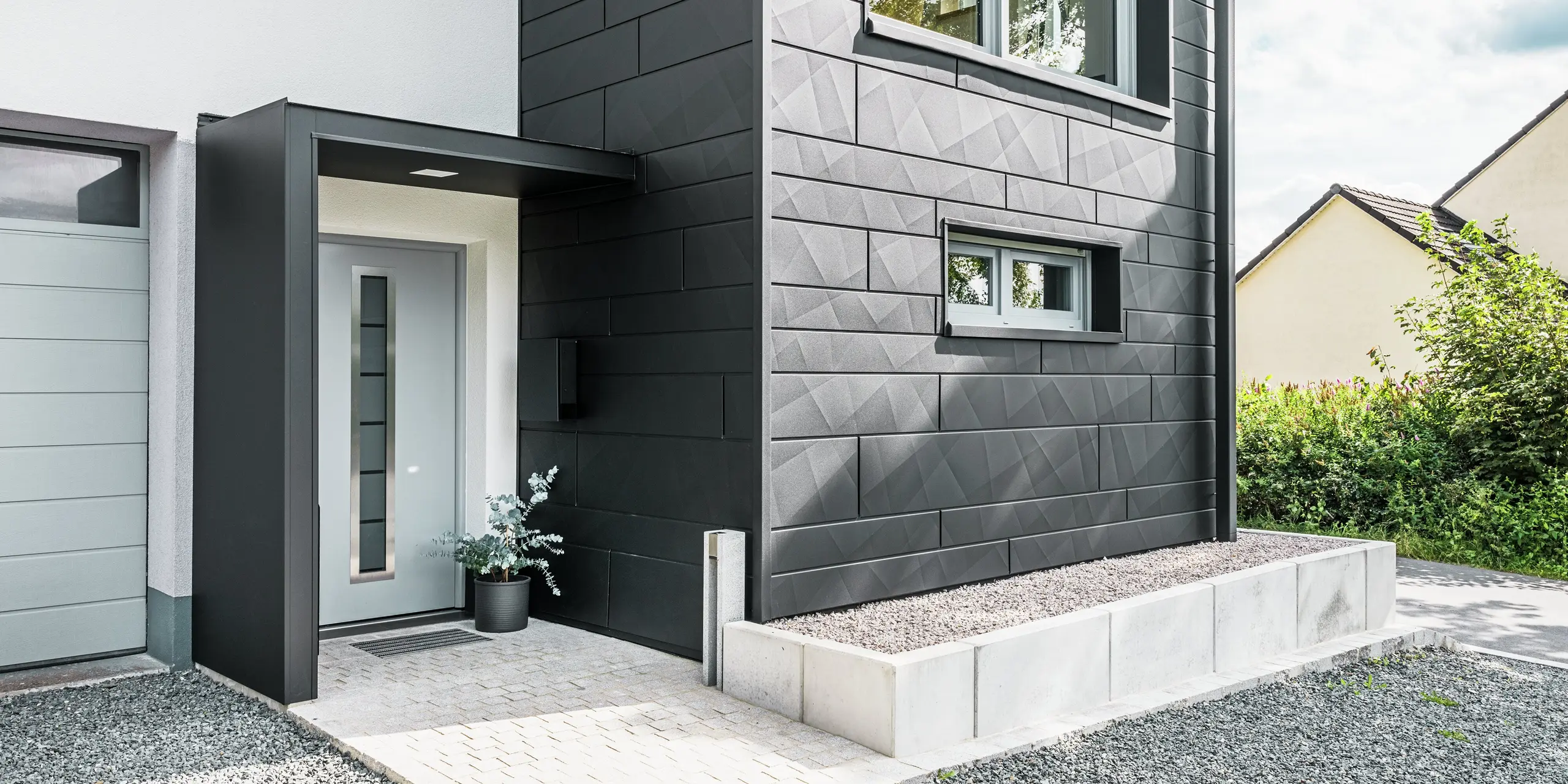L'élégante façade Siding.X PREFA en P.10 noir domine l'entrée de la maison individuelle modernisée de Teuschnitz. La façade en aluminium de haute qualité confère au bâtiment un caractère moderne et intemporel. Les panneaux de façade clairement structurés créent un design géométrique saisissant, tandis que les panneaux composites en aluminium noir encadrent la porte d'entrée et les fenêtres. Le tuyau de descente carré PREFA s'intègre harmonieusement au design sophistiqué et souligne l'impact fonctionnel et esthétique de la rénovation. L'allée menant à la maison est pavée de pavés et de gravier.