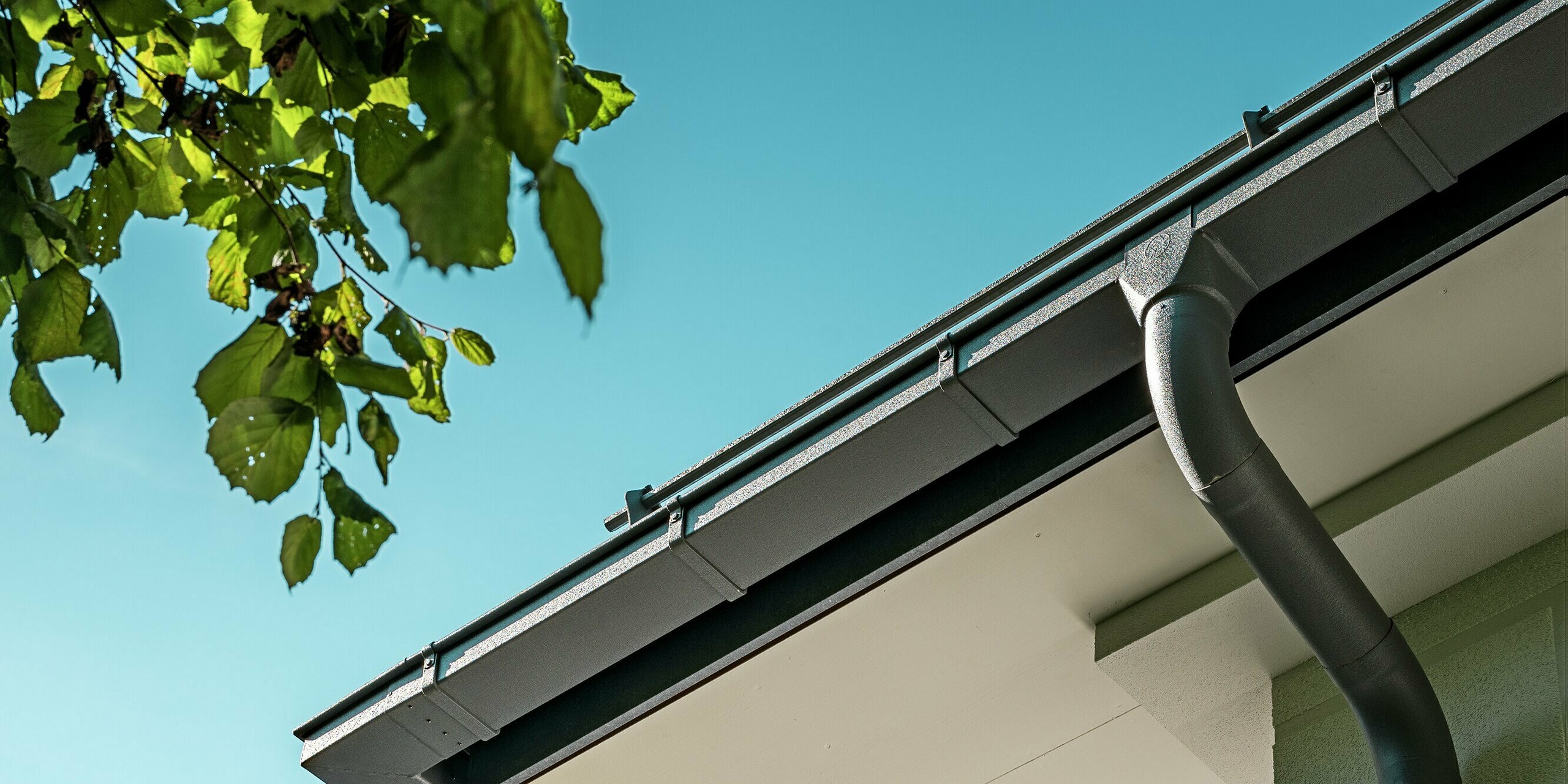 Gros plan sur le système de gouttière PREFA, avec sa gouttière carrée et son tuyau de descente, dans l'élégant coloris gris pierre P.10. Ce système en aluminium de haute qualité assure une évacuation efficace des eaux et s'intègre harmonieusement au design moderne de l'immeuble. La précision de la fabrication de la gouttière et son aspect contemporain soulignent la qualité esthétique et fonctionnelle de la toiture. Ses lignes épurées et l'attrait du matériau font de ce système d'évacuation des eaux pluviales un élément architectural majeur.
