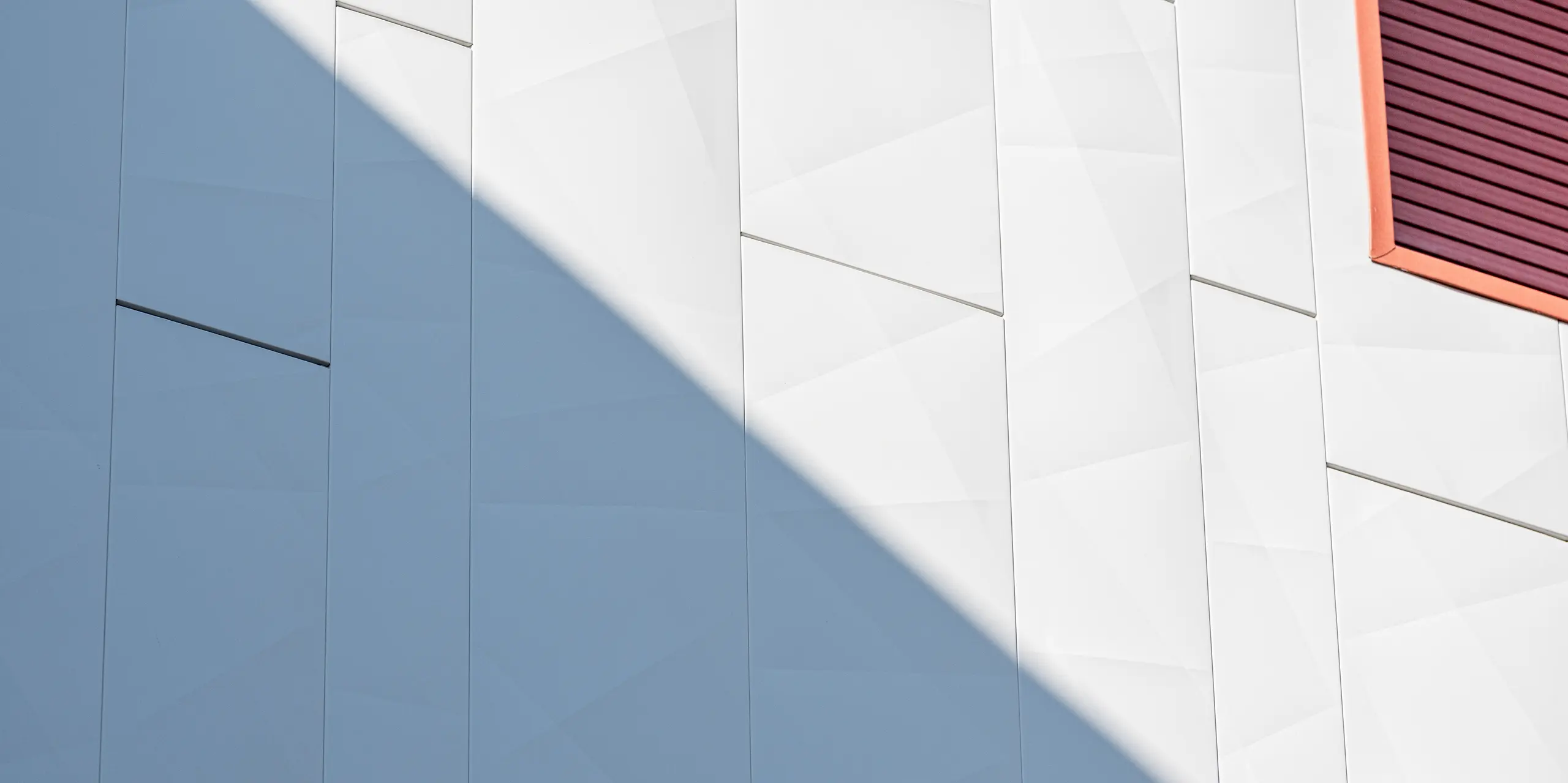 Détail de la façade rénovée du complexe résidentiel Alba B, revêtue de panneaux de façade en aluminium PREFA Siding.X : des panneaux grand format, posés verticalement et aux bords fins, créent une structure géométrique dynamique. Une ombre portée en diagonale souligne la surface métallique et crée un jeu d’ombre et de lumière saisissant. Sur le bord droit de l’image, un encadrement de fenêtre rehaussé de rouge apporte un contraste marqué. La façade en aluminium PREFA impressionne par sa haute résistance, sa durabilité et son esthétique soignée, ne nécessitant que peu d’entretien.