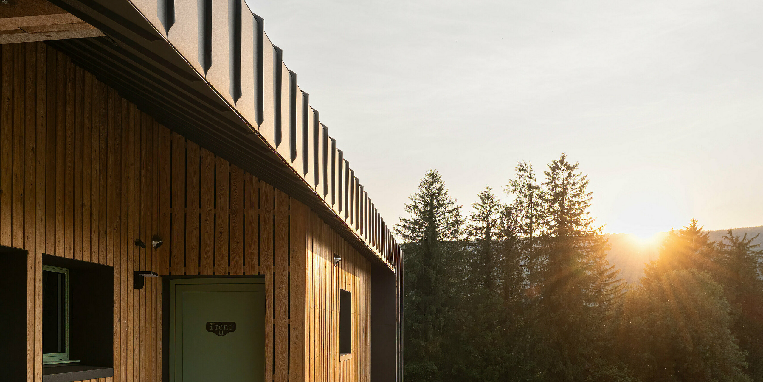 Vue latérale d'un bâtiment moderne avec une façade en bois et des éléments en aluminium au crépuscule. Les parties en aluminium de la maison sont en PREFALZ de couleur P.10 brun. Une rangée de chaises est installée à côté du mur devant le bâtiment. Il y a une balustrade métallique à l'étage supérieur. Le soleil se couche derrière les arbres à l’horizon, baignant la scène d’une lumière chaude.