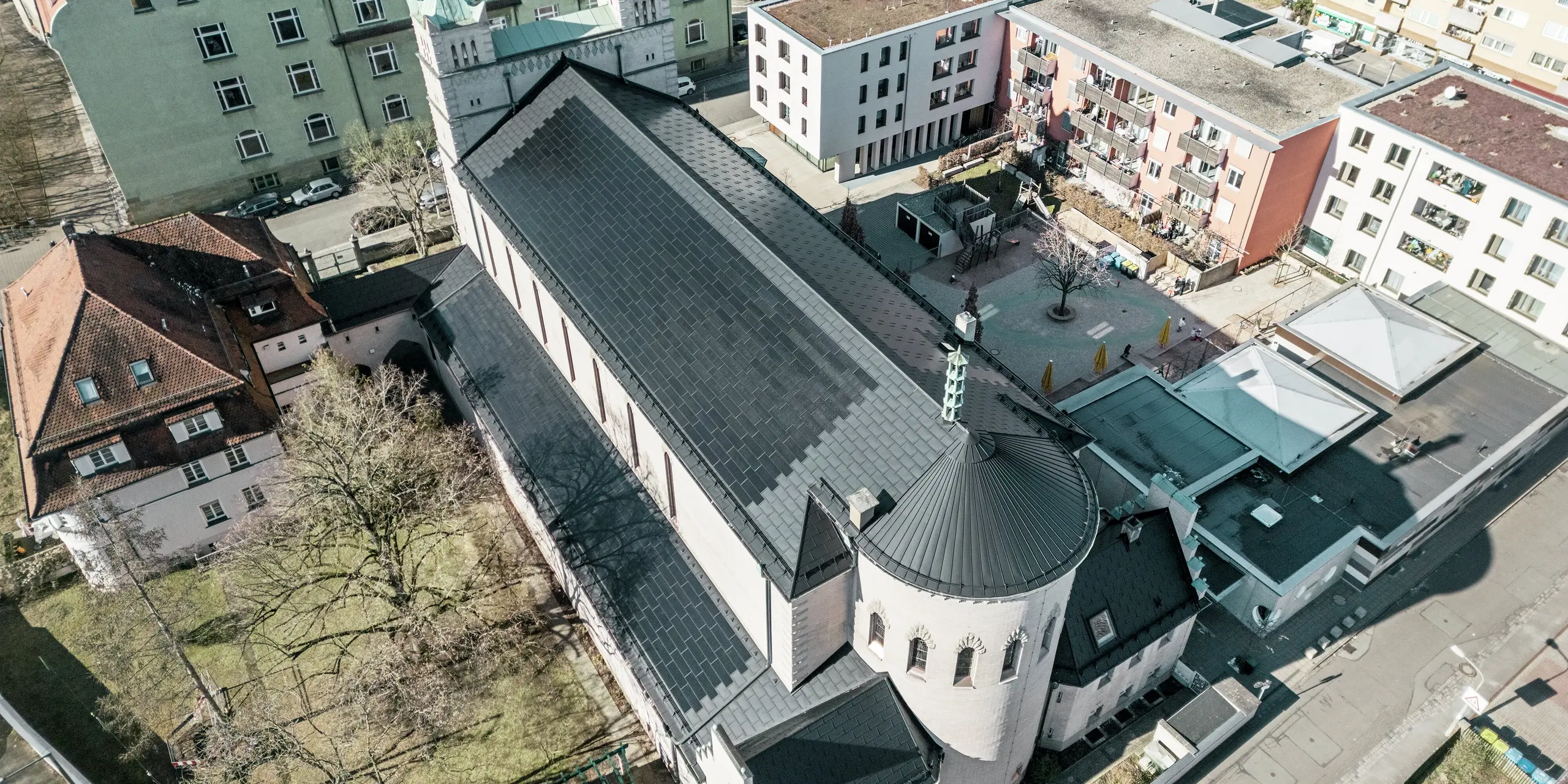 Vue aérienne de l'arrière de l'église Saint-Charles-Borromée de Nuremberg. Des tuiles solaires PREFA noires recouvrent le remarquable toit à pignon arrondi qui surplombe le corps principal de l'église, ainsi que celui du bâtiment adjacent. Les panneaux de toiture en aluminium avec modules photovoltaïques intégrés s'intègrent parfaitement à l'aspect historique de l'église. La vue sur l'environnement moderne, avec ses immeubles d'habitation adjacents et sa cour intérieure verdoyante, illustre l'alliance réussie entre préservation du patrimoine, architecture moderne et production d'énergie durable. Une solution idéale pour la préservation du patrimoine en milieu urbain.