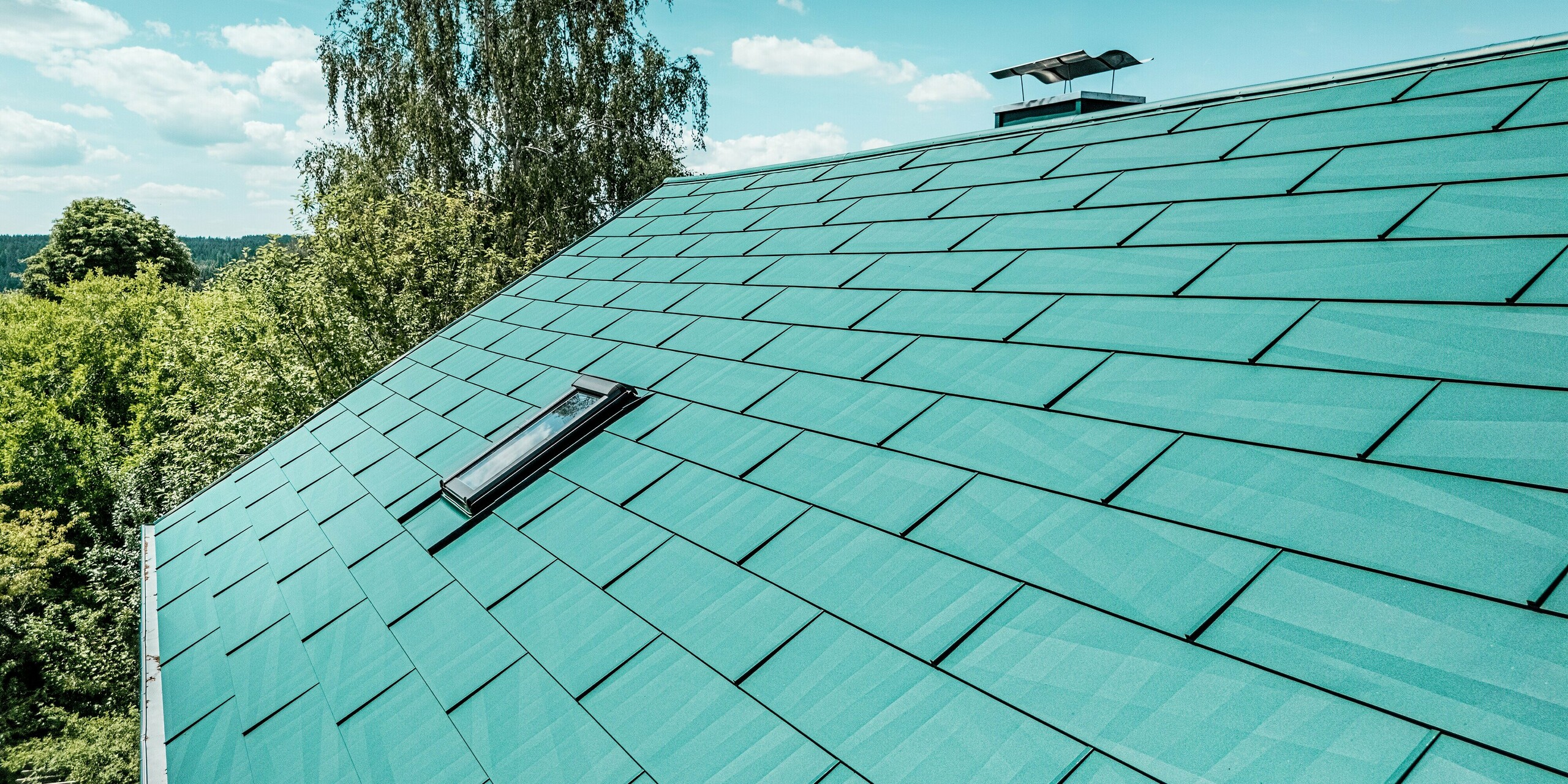 Détail de la toiture rénovée d'une maison individuelle à Schwarzenbach-Martinlamitz, réalisée avec des panneaux PREFA FX.12 couleur Vert Mousse P.10. L'image met en valeur la surface structurée en aluminium aux bords irréguliers et modernes, qui reflète élégamment la lumière du soleil. La remarquable verrière et la cheminée élégamment revêtue ajoutent une touche esthétique. Durables, les panneaux PREFA offrent une protection optimale contre les intempéries, ne nécessitent aucun entretien, résistent aux tempêtes et sont exceptionnellement légers – la solution idéale pour des rénovations de toiture haut de gamme à vocation architecturale.