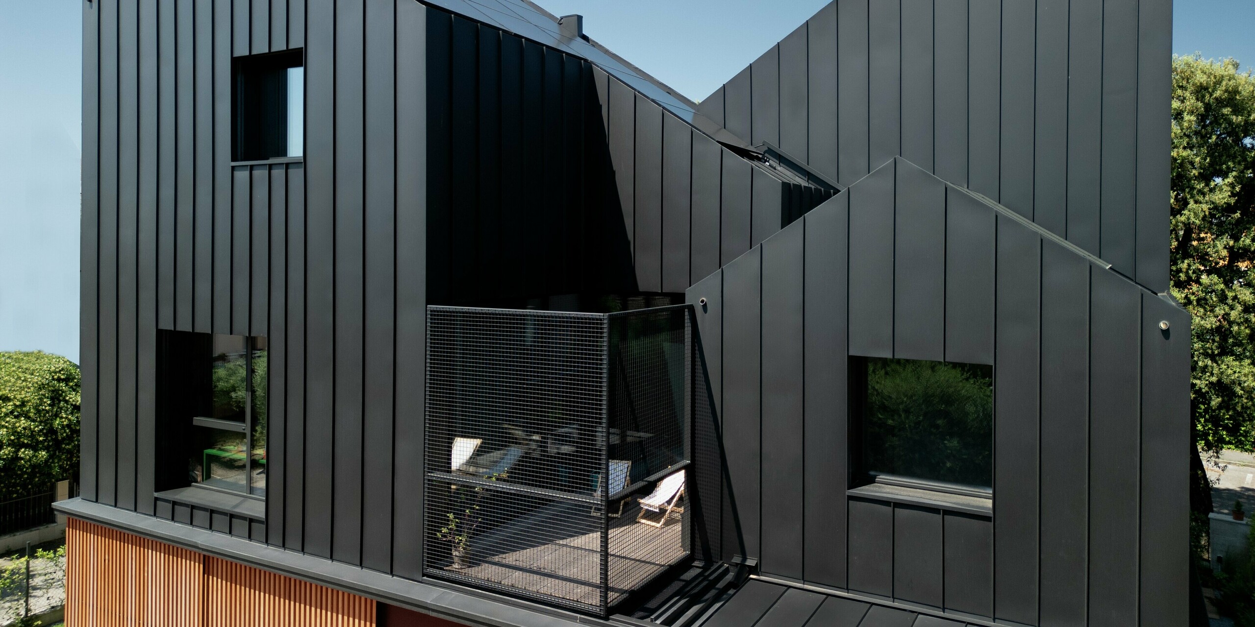 Vue d'ensemble de l'étage supérieur d'une maison jumelée moderne à Cattolica : façade et toiture en aluminium PREFALZ gris noir, associées à un bardage en bois chaleureux. La pureté des lignes et la qualité de la surface PREFA garantissent durabilité, protection contre la corrosion et un design élégant et intemporel sous un ciel bleu.