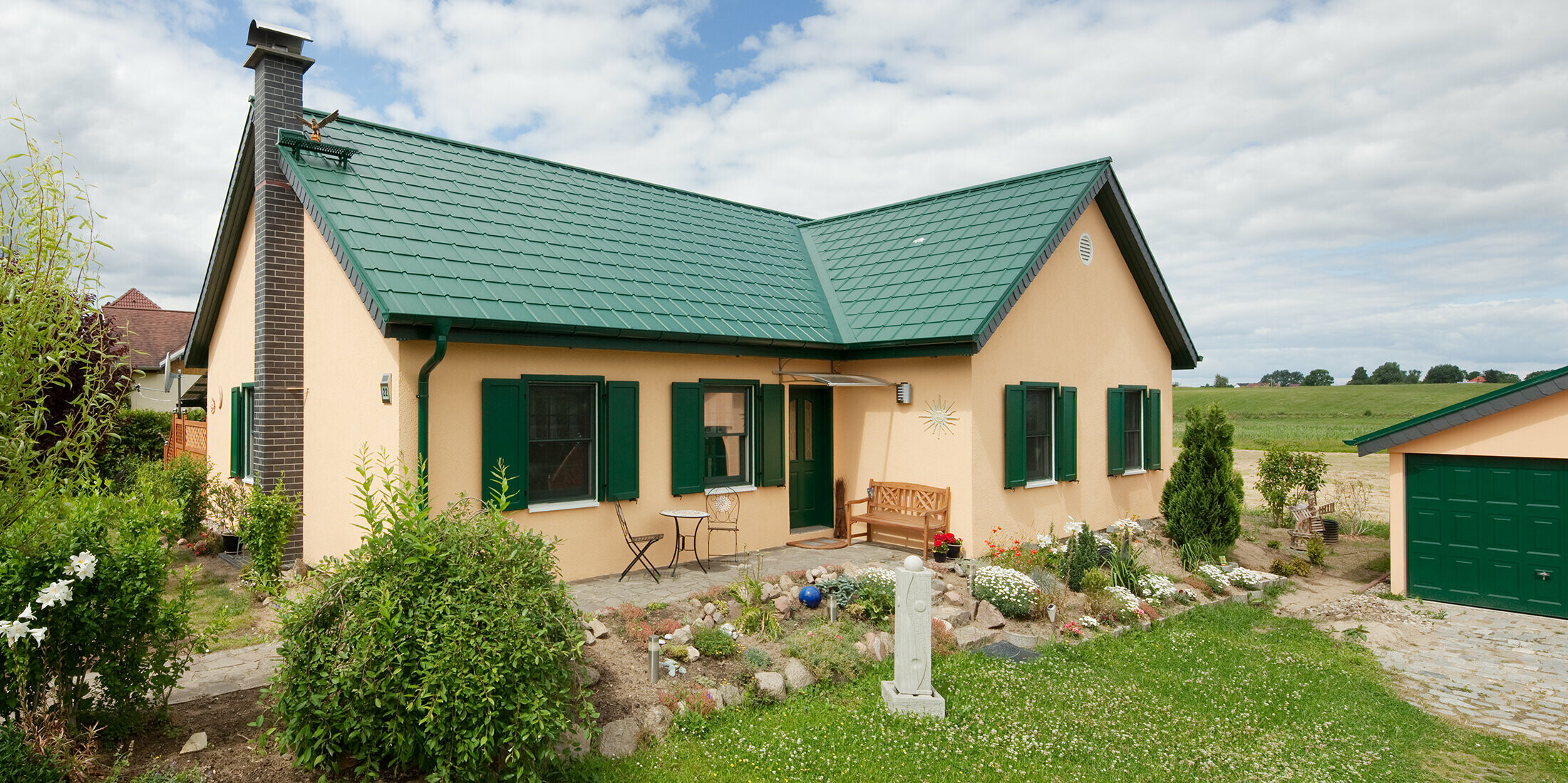 Toit en bâtière élargi ; toit en bâtière en forme de L ; avec la tuile PREFA en P.10 vert mousse ; jolie petite maison de campagne avec des volets verts assortis à la toiture PREFA.