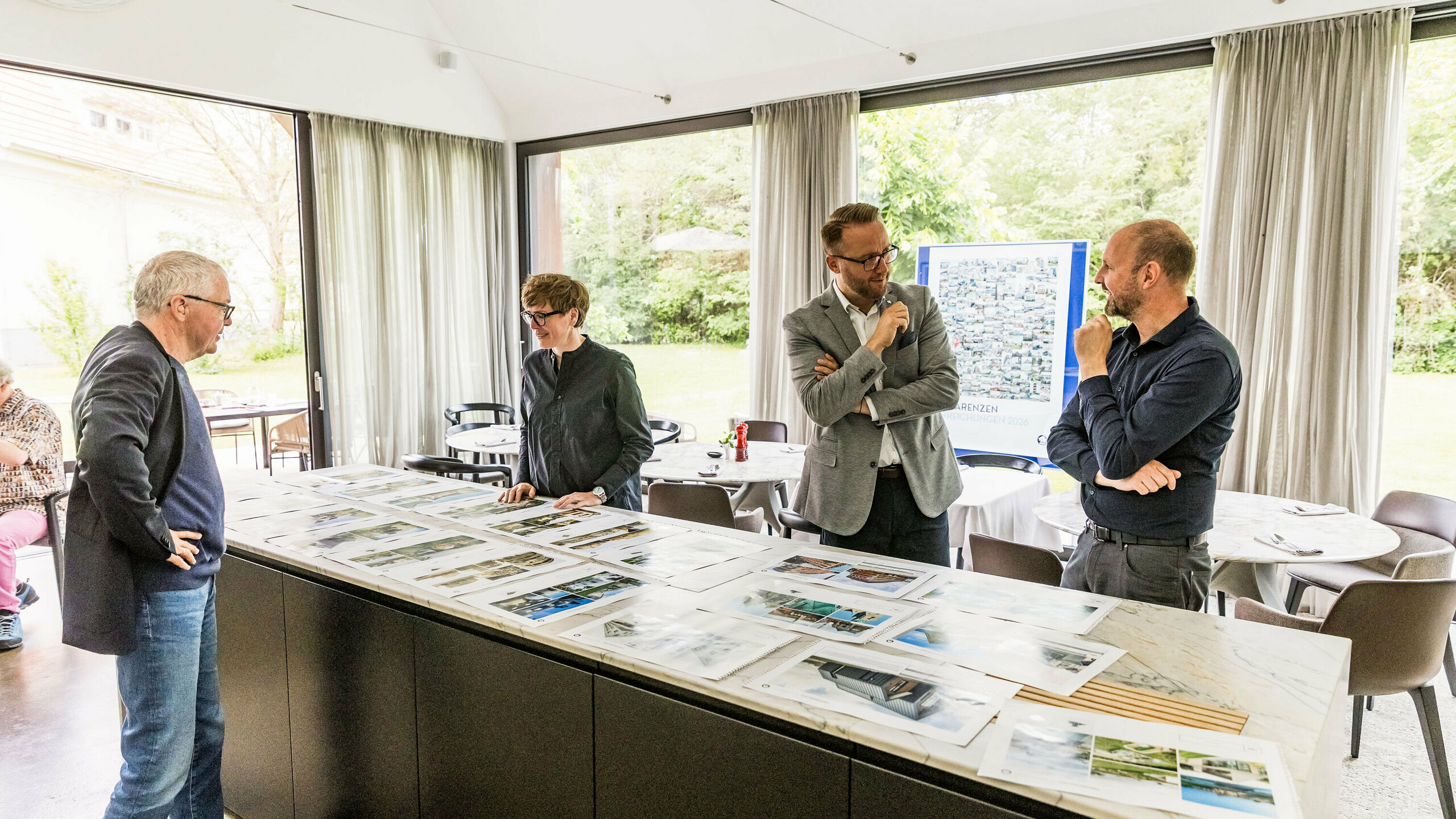 La sélection finale des points forts architecturaux PREFARENZEN pour 2026 est présentée en grandes images devant une partie du jury de haut niveau. Les experts examinent attentivement les projets architecturaux soumis en termes de force d'innovation, de qualité de conception et d'utilisation des matériaux PREFA. La réunion dans la maison vigneronne inondée de lumière à Seewinkel dans le Burgenland marque la conclusion du processus intensif du jury pour le livre d'architecture PREFARENZEN et le calendrier 2026.