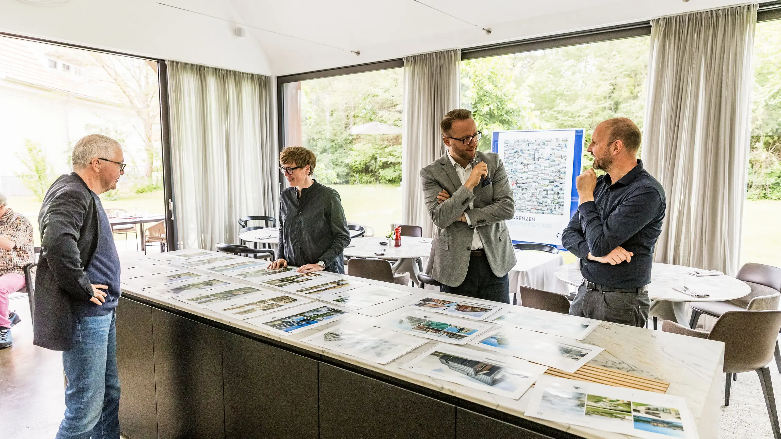 La sélection finale des points forts architecturaux PREFARENZEN pour 2026 est présentée en grandes images devant une partie du jury de haut niveau. Les experts examinent attentivement les projets architecturaux soumis en termes de force d'innovation, de qualité de conception et d'utilisation des matériaux PREFA. La réunion dans la maison vigneronne inondée de lumière à Seewinkel dans le Burgenland marque la conclusion du processus intensif du jury pour le livre d'architecture PREFARENZEN et le calendrier 2026.