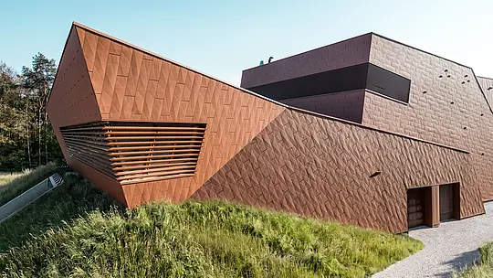 La remarquable station de pompage d'eau potable WML de Lottum se distingue par sa structure aux plis dynamiques et son faîte caractéristique qui attire immédiatement le regard. La façade en aluminium PREFA Siding.X, à l'aspect chaleureux rappelant la rouille, séduit par sa texture facettée et vibrante et par le jeu subtil d'ombre et de lumière. Des persiennes horizontales soulignent la profondeur de l'architecture, tandis que le bâtiment s'intègre harmonieusement à son environnement naturel. Légers, durables et recyclables, les panneaux en aluminium PREFA offrent une résistance maximale aux intempéries et une enveloppe de bâtiment durable et nécessitant peu d'entretien.