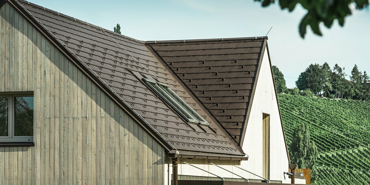 Cette maison individuelle avec deux toits à pignon est recouverte de bardeaux de toiture PREFA en brun noisette. L'écoulement des eaux de pluie se fait par la gouttière carrée PREFA. La façade est revêtue de bois patiné.